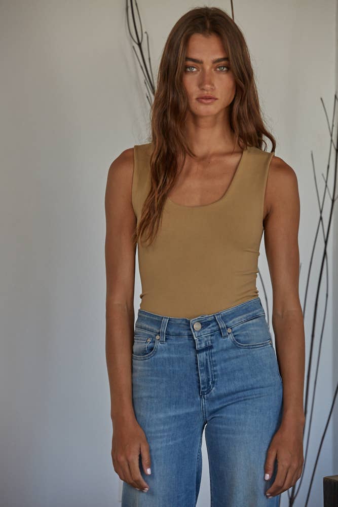 A woman with long brown hair wears a By Together Double Layered Seamless Top and high-waisted blue jeans, standing indoors against a light wall with decorative branches in the background.