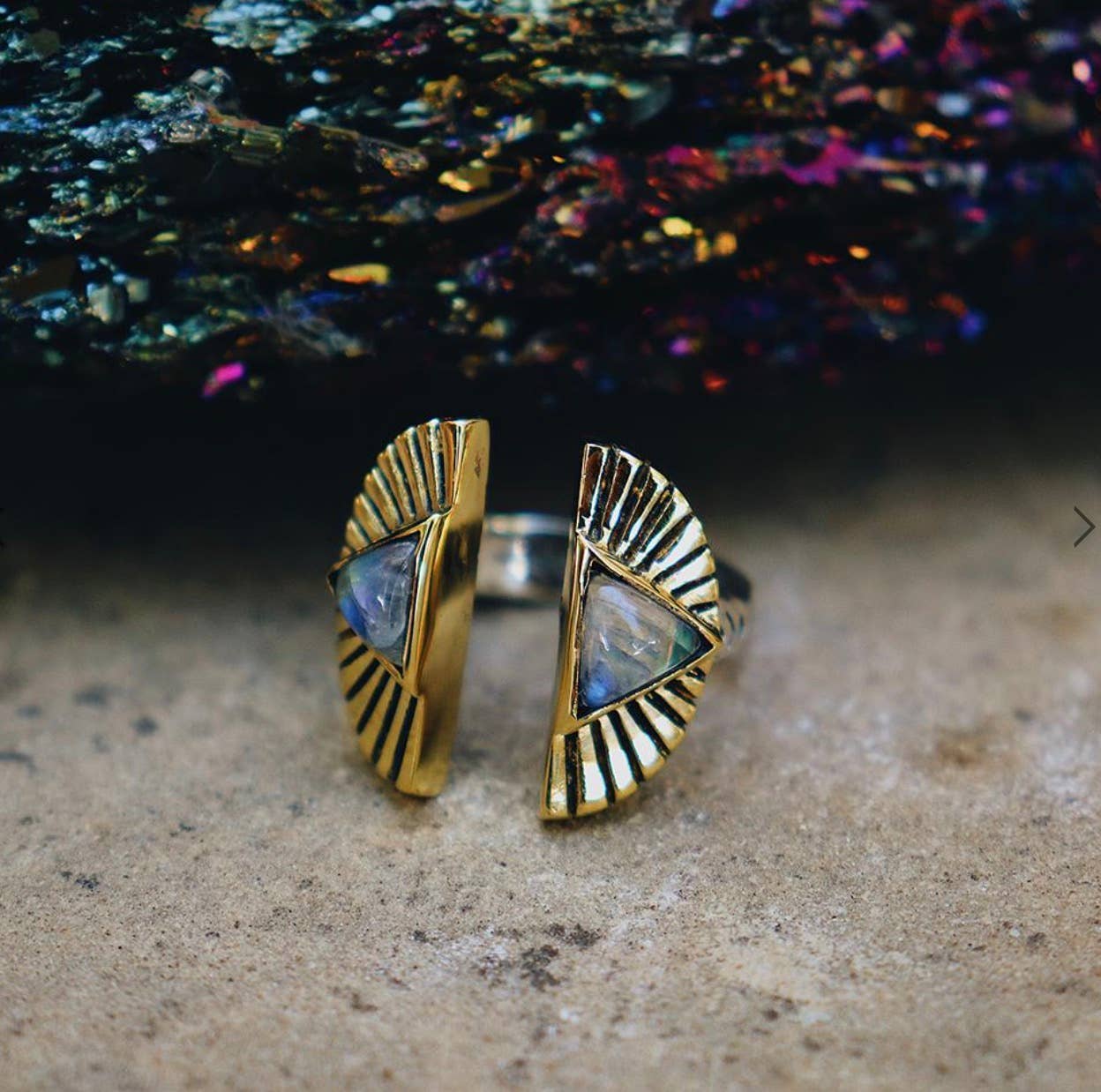 The Shape Shift Ring by bahgsu jewels is a gold adjustable spiritual ring with two textured semicircular ends and triangular gemstones, shown on stone with a colorful, iridescent background.