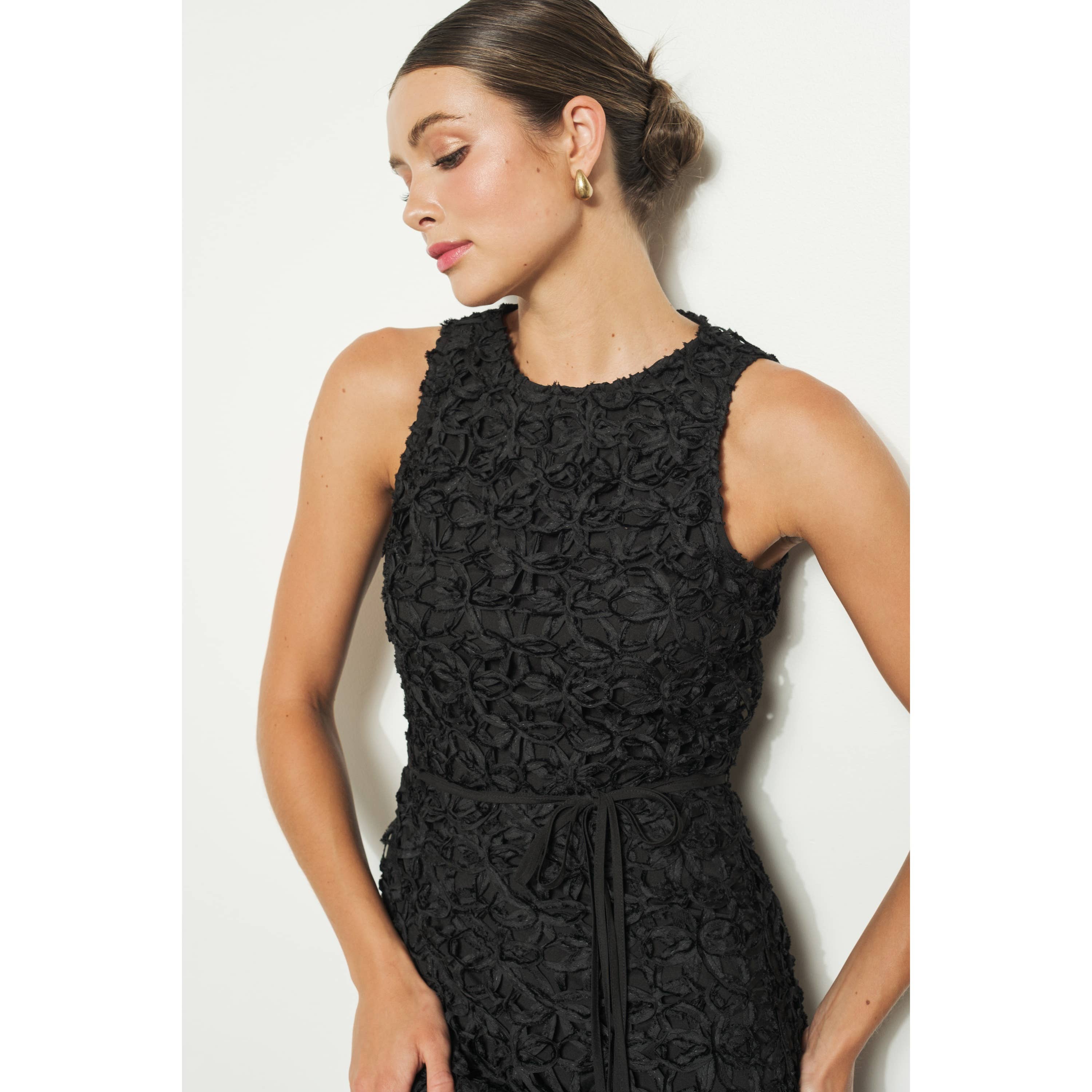 A woman models the INA Sleeveless Crochet Lace Overlay Midi Dress with a tied waist, standing against a white background. Her hair is styled in a neat bun, and she wears gold hoop earrings while gazing down and to the side.