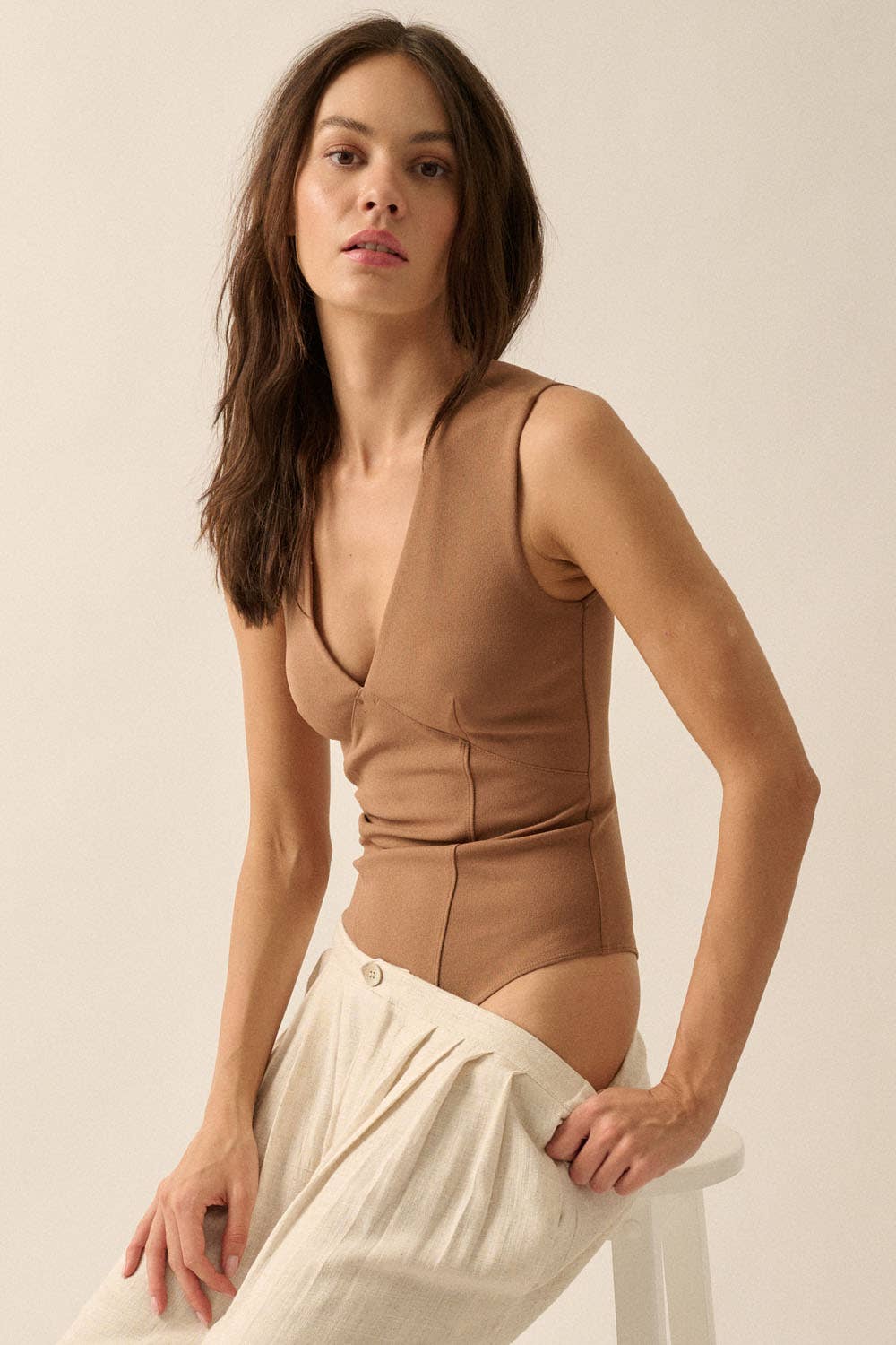 Woman wearing a brown Essential Trends Crepe V-Neck Bodysuit seated on a white stool with light pants.