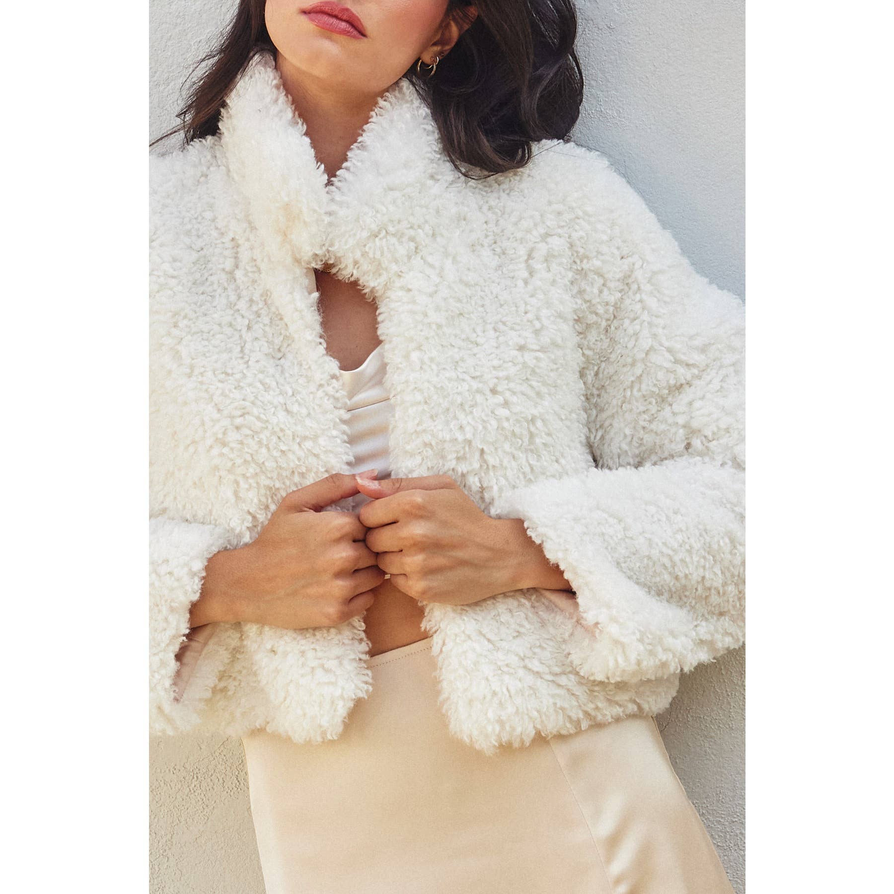 A woman stands against a light background, touching the Soft Teddy Open Front Cropped Jacket by Dress Forum, layered over a light satin dress. Her face is partially out of frame, highlighting the fluffy texture of the cozy cropped jacket.