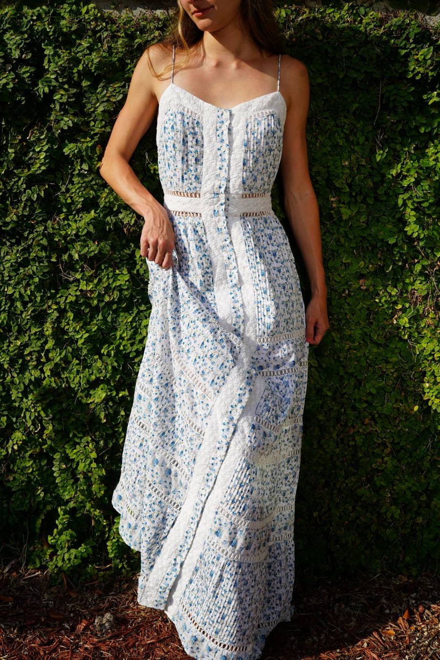 Woman wearing a light, floral-patterned Holly Dress with delicate straps, standing against a green leafy background