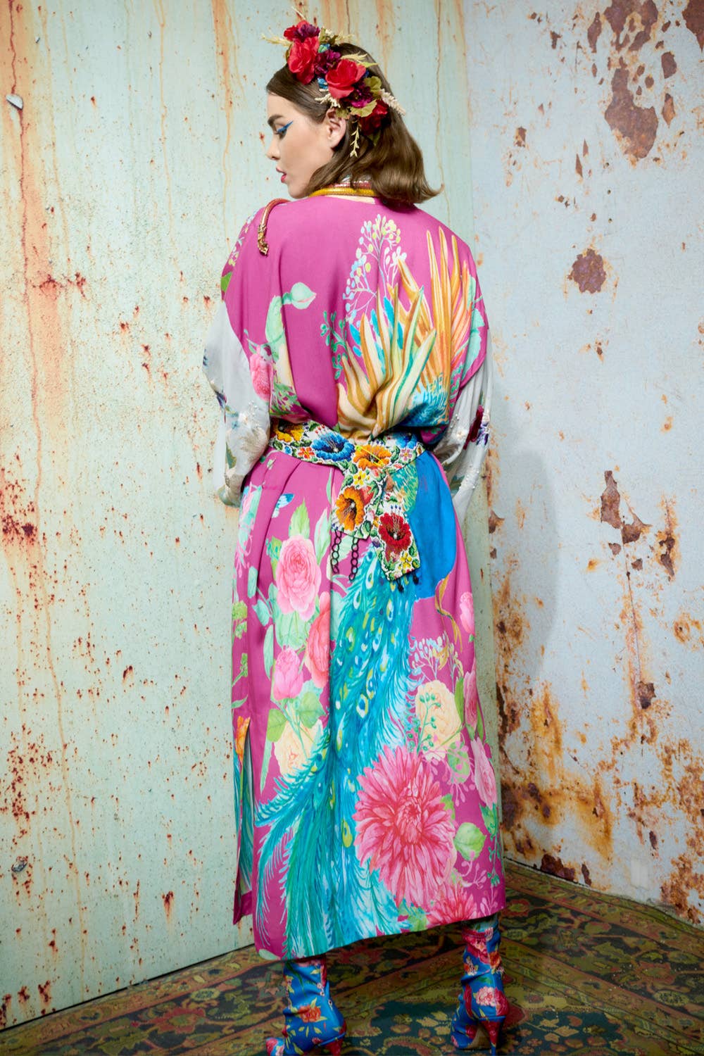 A woman faces away from the camera in Aratta's Marie Antoinette Kimono, featuring embroidered floral sleeves. With flowers in her hair and matching boots, she stands against a distressed wall and patterned floor.