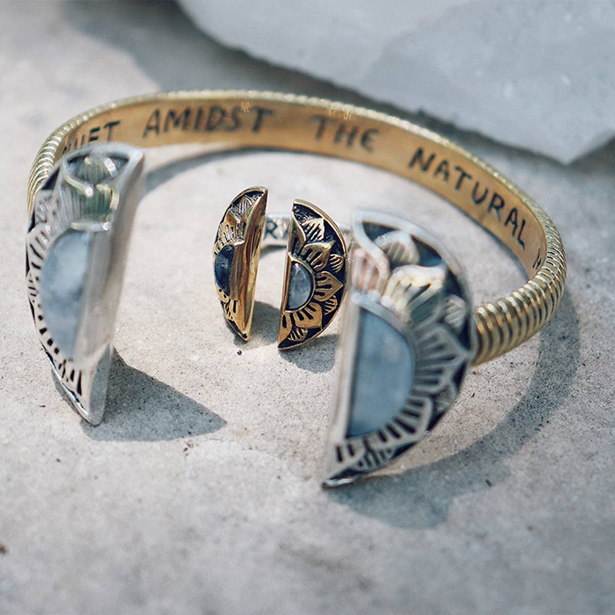 The bahgsu jewels Natural Mystic Ring - Moonstone and a matching silver and gold cuff bracelet with moon phase designs rest on a stone surface. The bracelets interior reads AMIDST THE NATURAL.