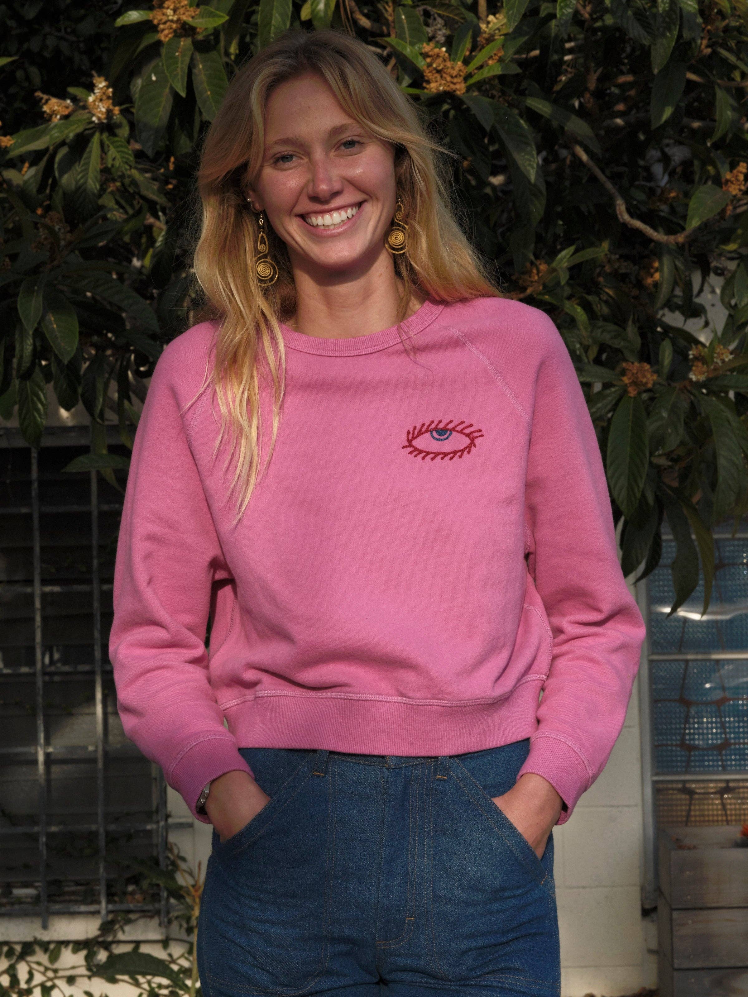 A smiling woman with long blonde hair wears the Mollusk Visions Crew, a relaxed fit 100% cotton sweatshirt featuring an eye design, paired with blue jeans as she stands outdoors by leafy green foliage and a window.