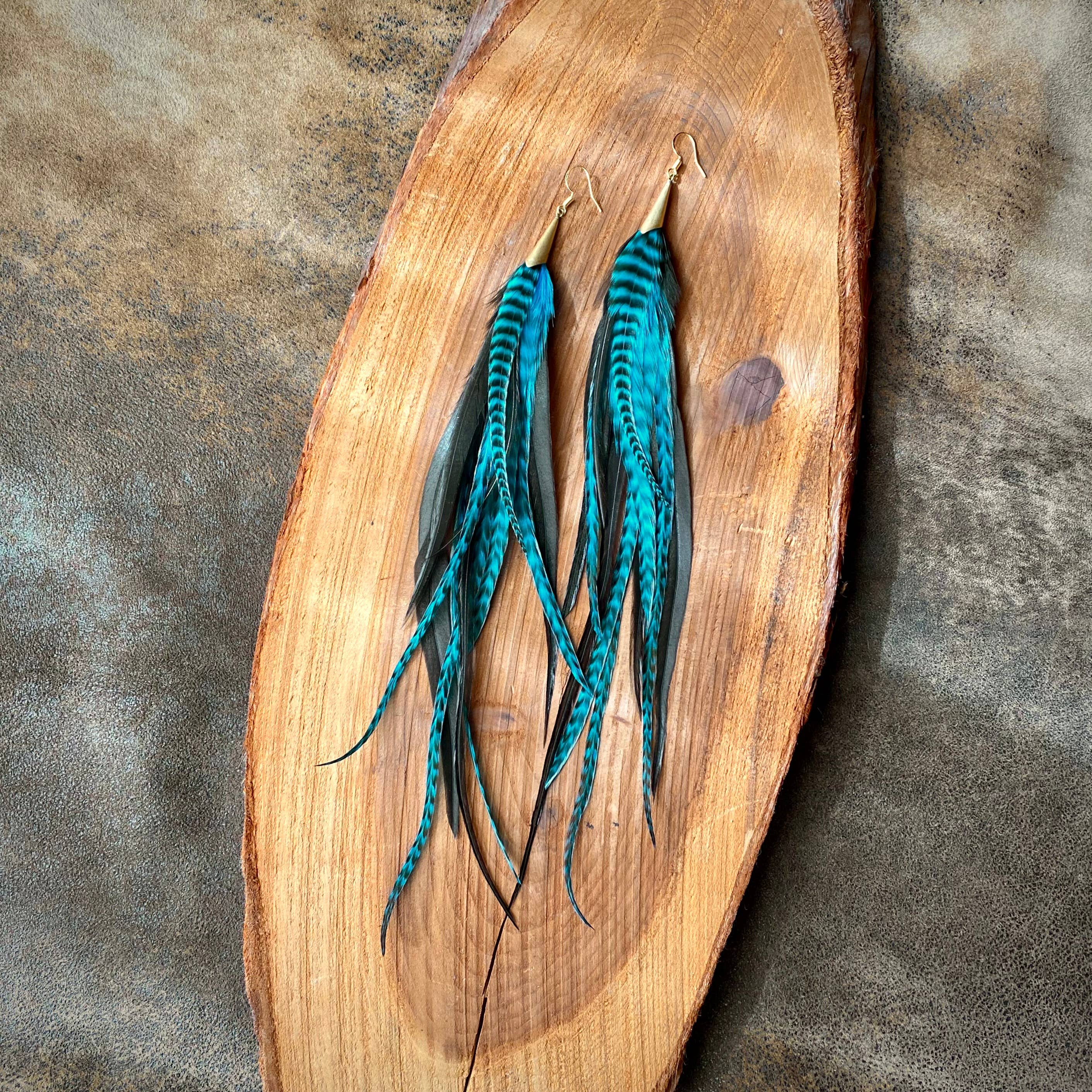 The ASTALI Long Feather & Cone Earrings - Turquoise Grizzly & Black/Gold, featuring sustainably sourced feathers and gold hooks, are displayed on an oval wooden tray against a textured gray and brown background.