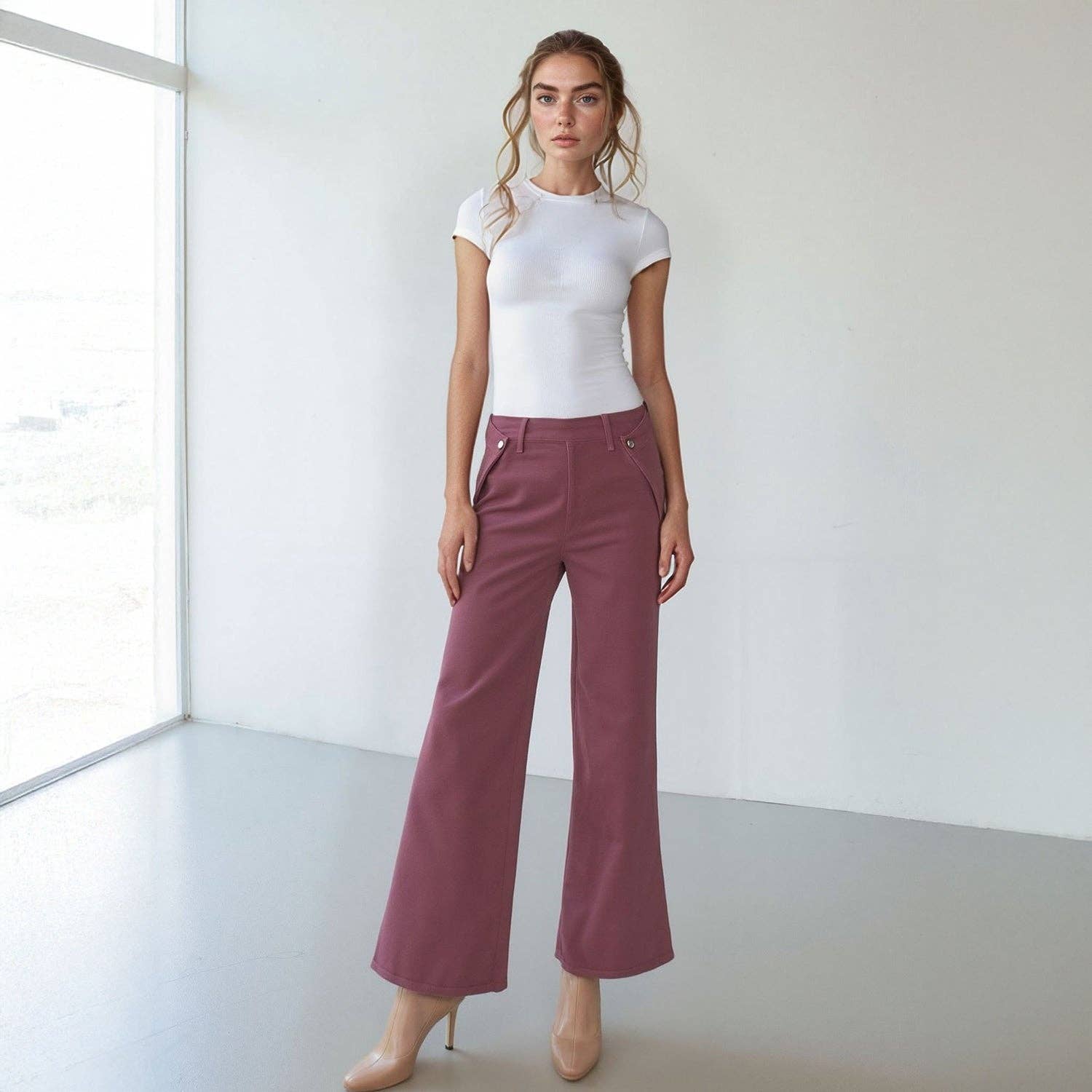 A woman stands indoors wearing a fitted white short-sleeve top, nude heels, and Noend Denim’s Dina High Waisted Wide Corduroy pants in Deep Merlot, her hands by her sides and a neutral expression.