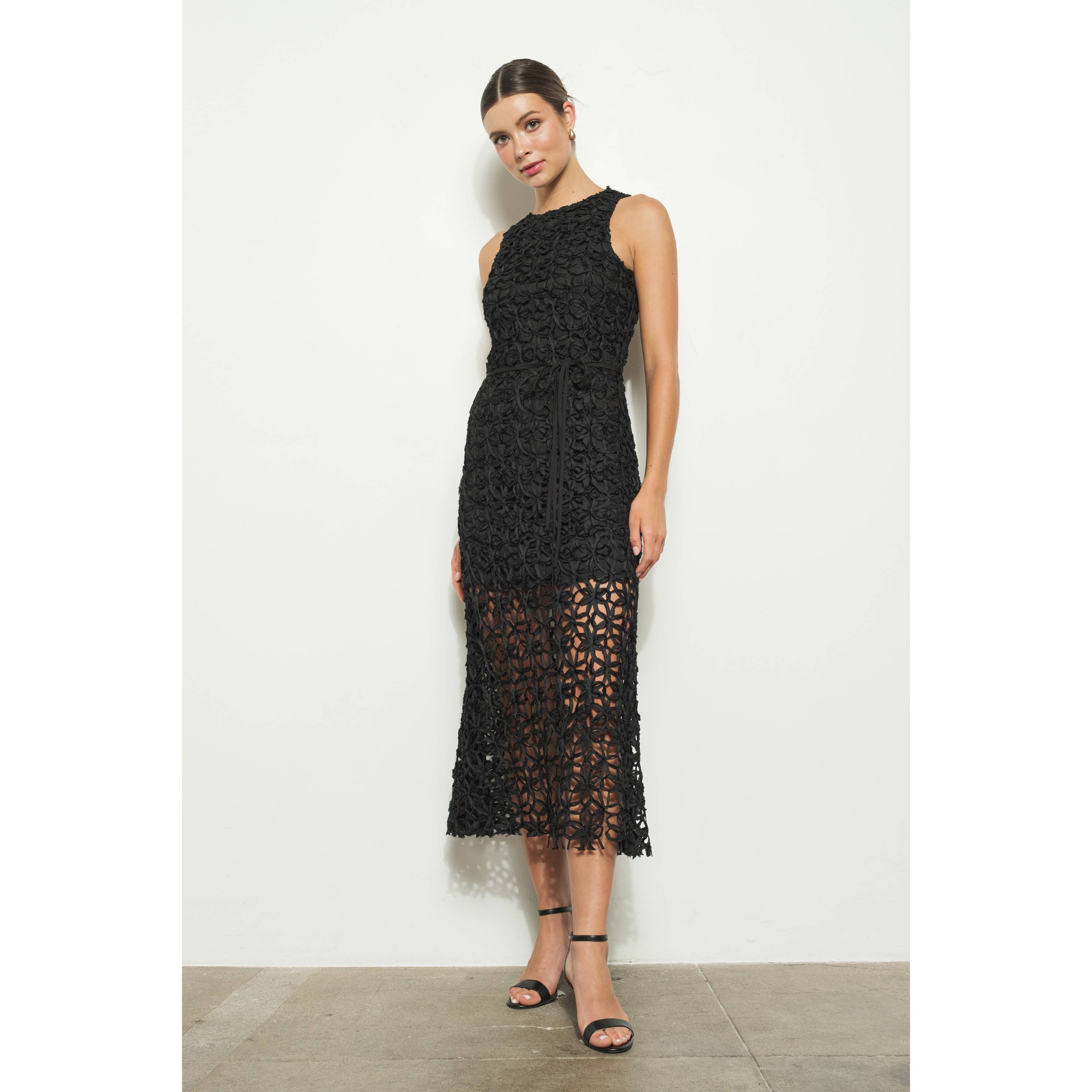 A woman stands against a white wall wearing the INA Sleeveless Crochet Lace Overlay Midi Dress with a high neckline and sheer panels. Her hair is pulled back, and she pairs the dress with black strappy sandals.