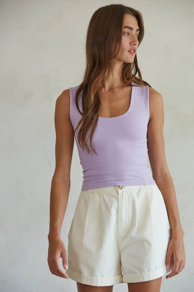 A woman with long brown hair wears the By Together Double Layered Seamless Top in lavender and high-waisted white shorts, standing against a light background and looking to her left.