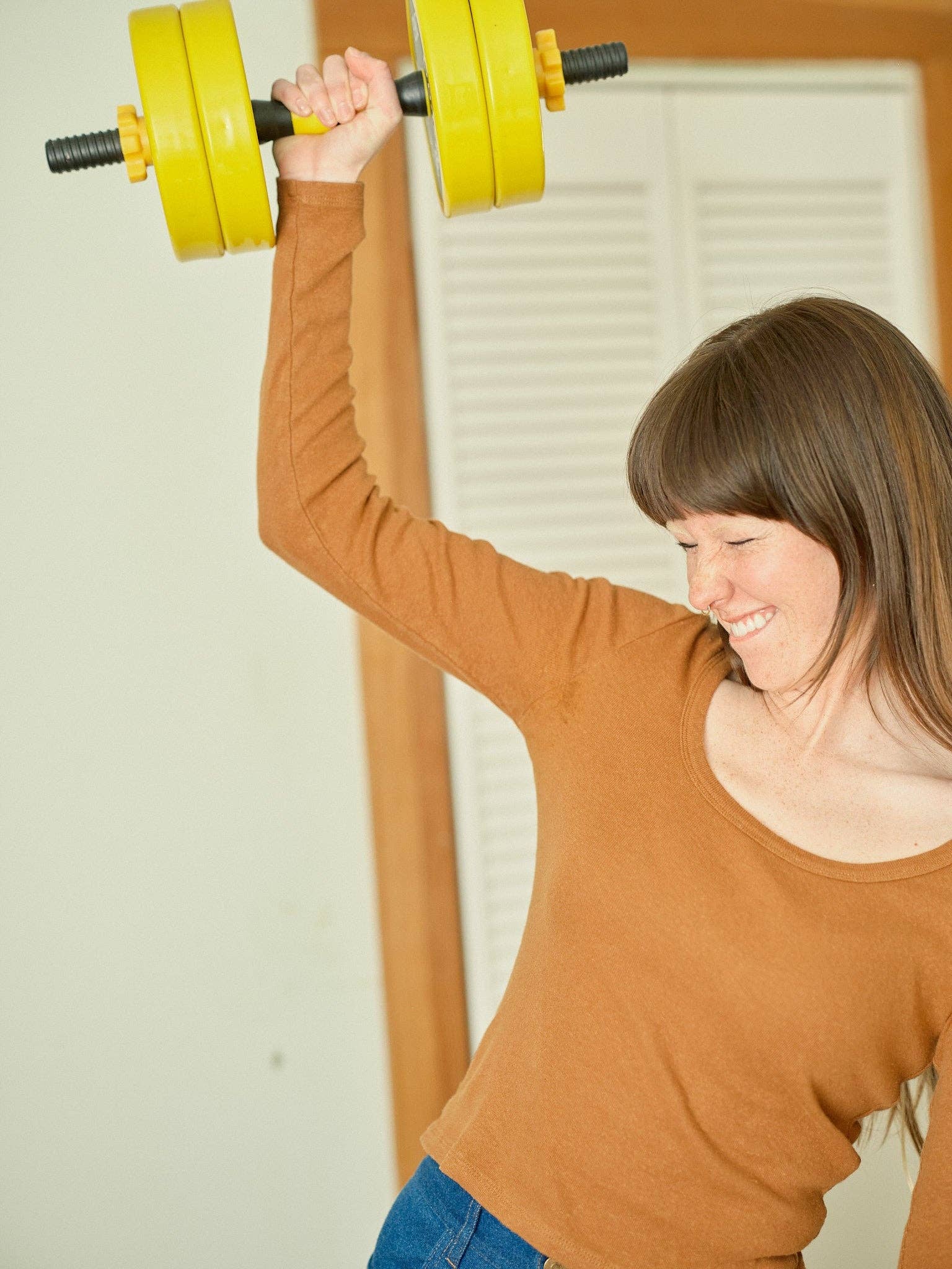 A woman with long brown hair wears the Mollusk Long Sleeve Rib Tee and smiles as she lifts a yellow dumbbell overhead with one arm.