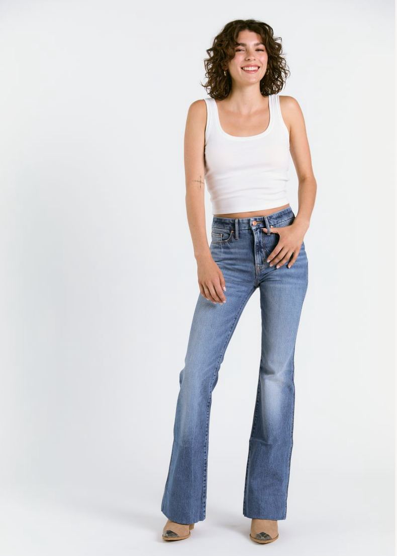 Smiling person with curly brown hair wears a white sleeveless crop top and Porter Blue Apparels WANDERER 5 POCKET FLARE IN MABEL WASH jeans, plus beige shoes, standing against a plain white background.