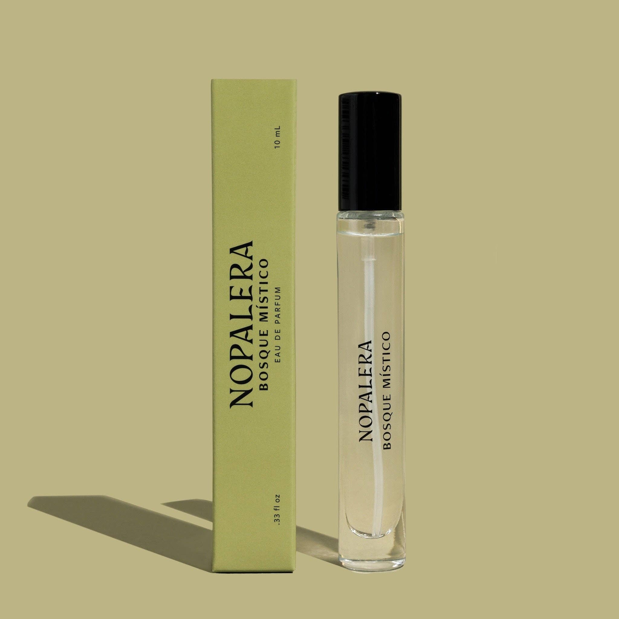 A clear glass Bosque Místico Eau de Parfum – 10mL Travel Spray by Nopalera stands beside its light green box on a beige background, both casting a soft left shadow, evoking the scent of a mystical forest.