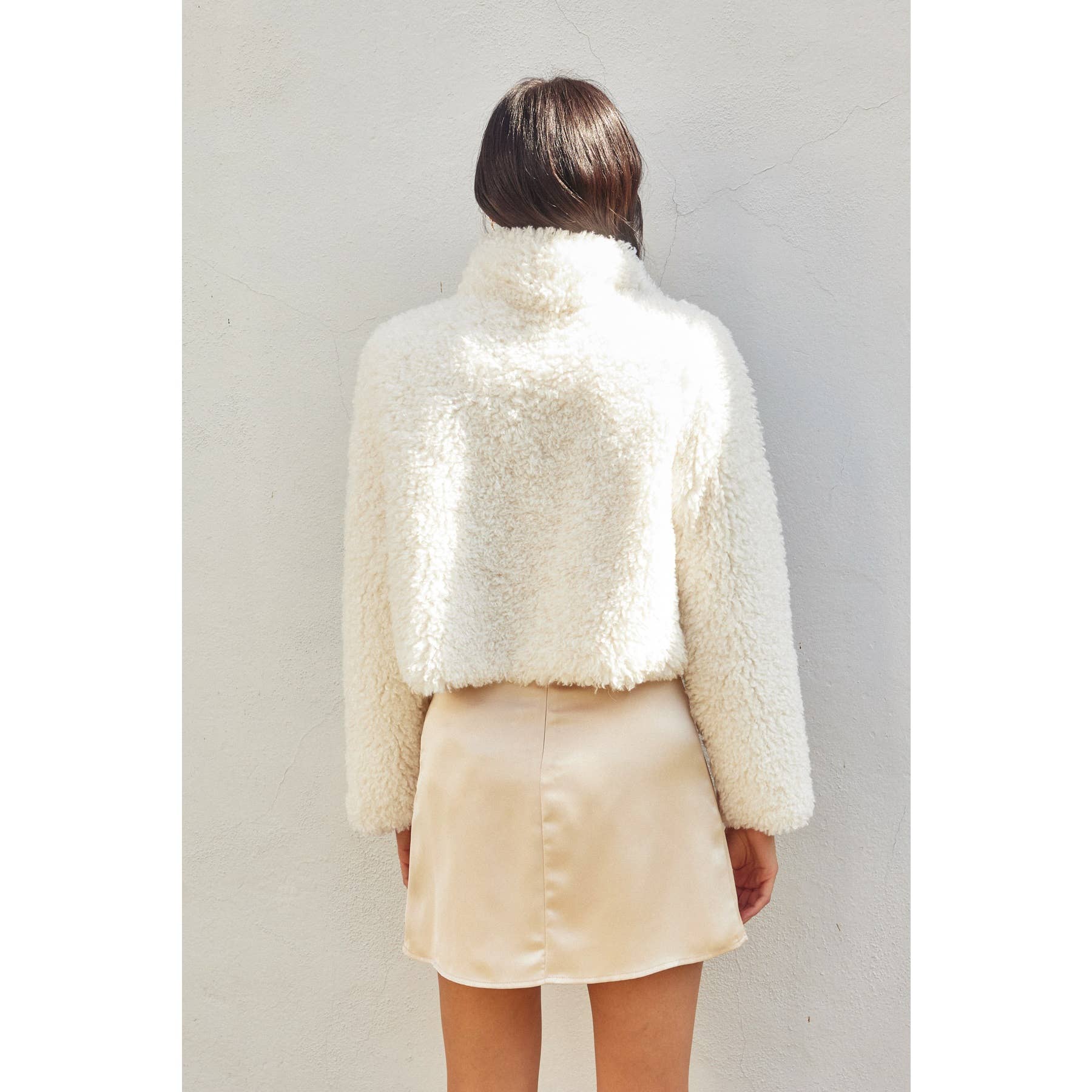 A woman with loose brown hair faces a white wall, wearing the Dress Forum Soft Teddy Open Front Cropped Jacket in cream and a pale beige satin skirt, photographed from behind.