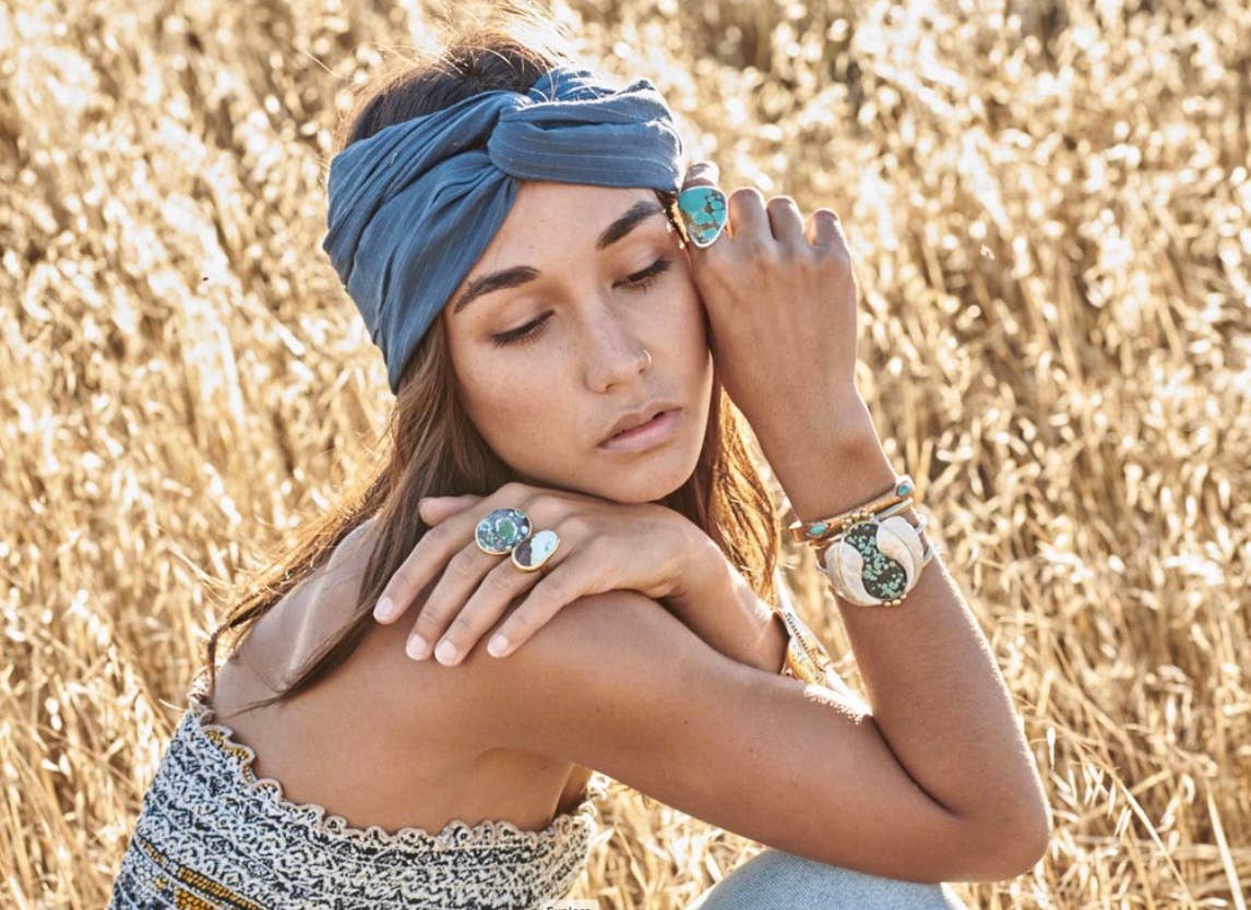 A woman in a blue headband and patterned top sits in a golden field, eyes closed. She wears large statement rings and the bahgsu jewels Medicine Wheel Cuff, a limited edition hand-carved piece with turquoise inlay, on her wrist.