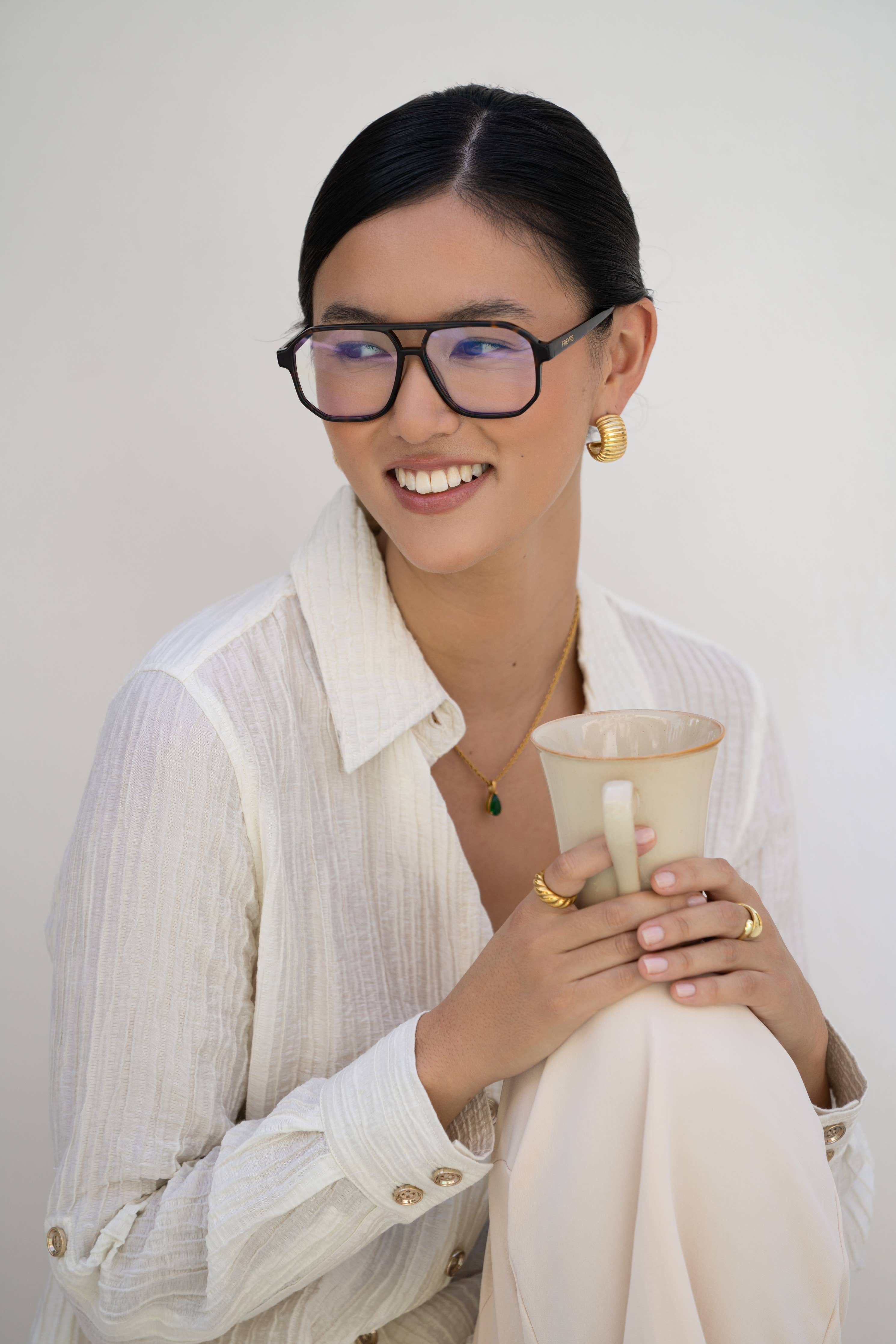 A woman with dark hair and gold jewelry smiles while holding a light cup, wearing FREYRS Eyewear Billie Blue Light Readers reading glasses with handmade acetate frames, dressed in a textured white blouse against a plain light background.