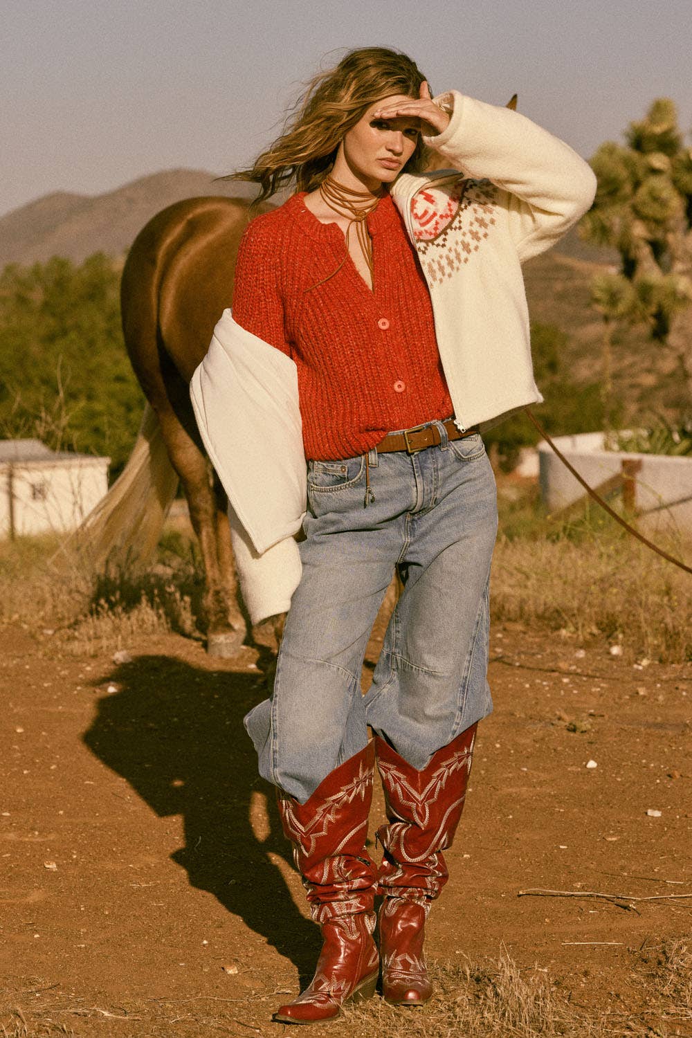 A woman wearing the Promesa USA Confetti Button-Front Cardigan Sweater, baggy jeans, and red cowboy boots stands outdoors shielding her eyes from the sun as a brown horse grazes nearby amid trees and hills in the background.
