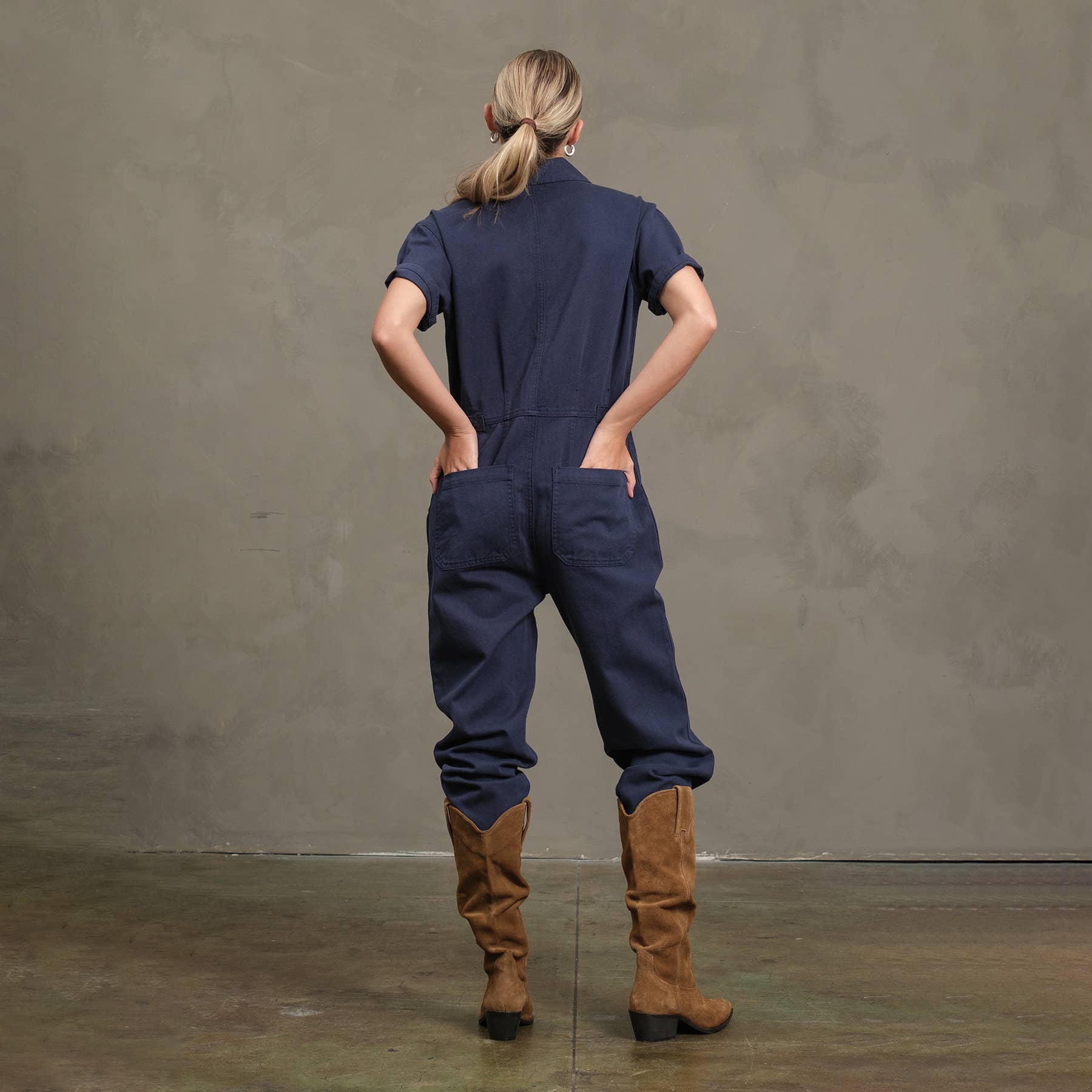 A person stands with hands in the back pockets of a dark blue RAISONNEL Field Jumpsuit (RJP1001) with rolled-up legs, paired with tall brown suede boots, against a plain neutral wall and floor.