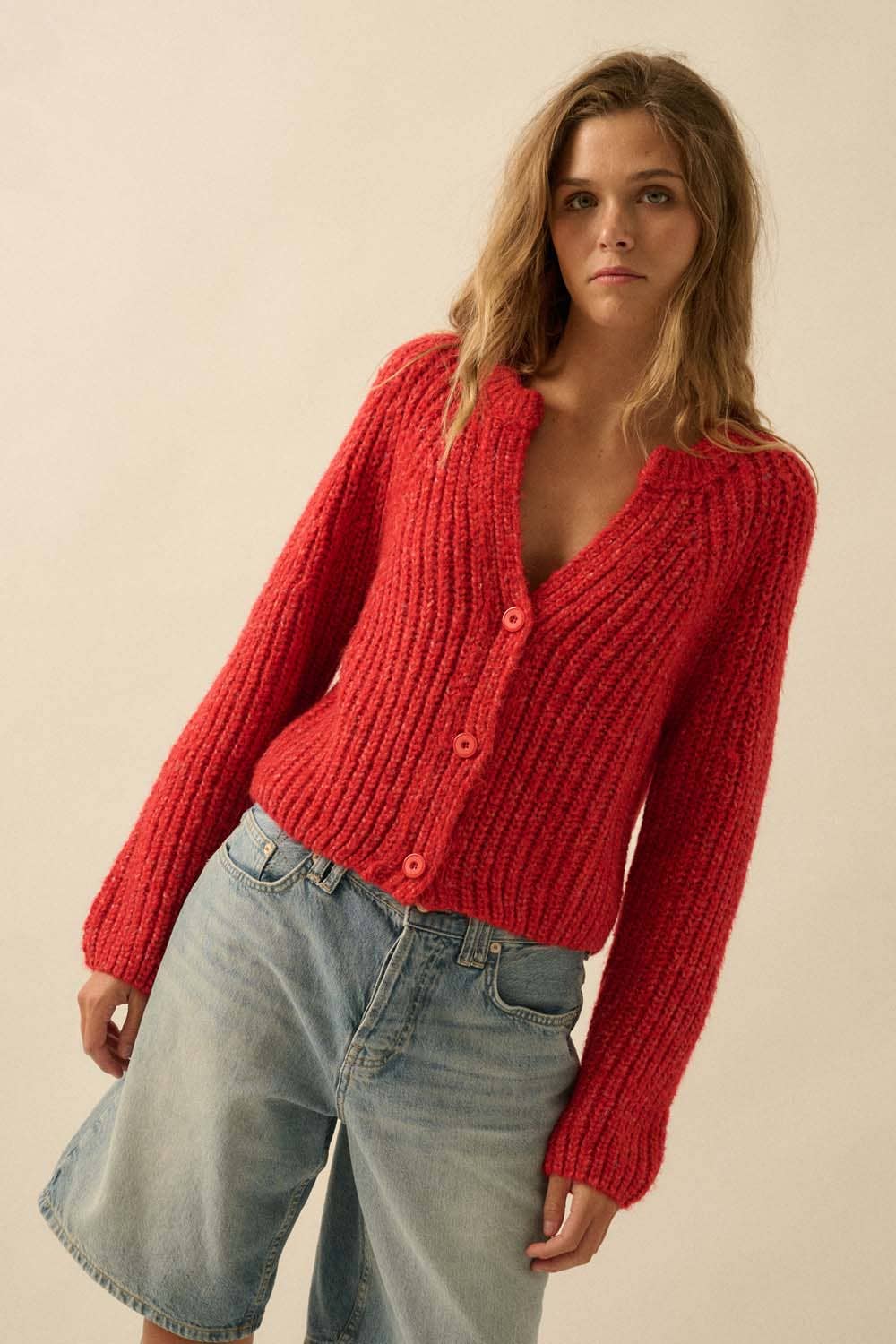 A woman with wavy blonde hair models the Promesa USA Confetti Button-Front Cardigan Sweater with large buttons and light blue denim shorts, standing against a plain beige background.