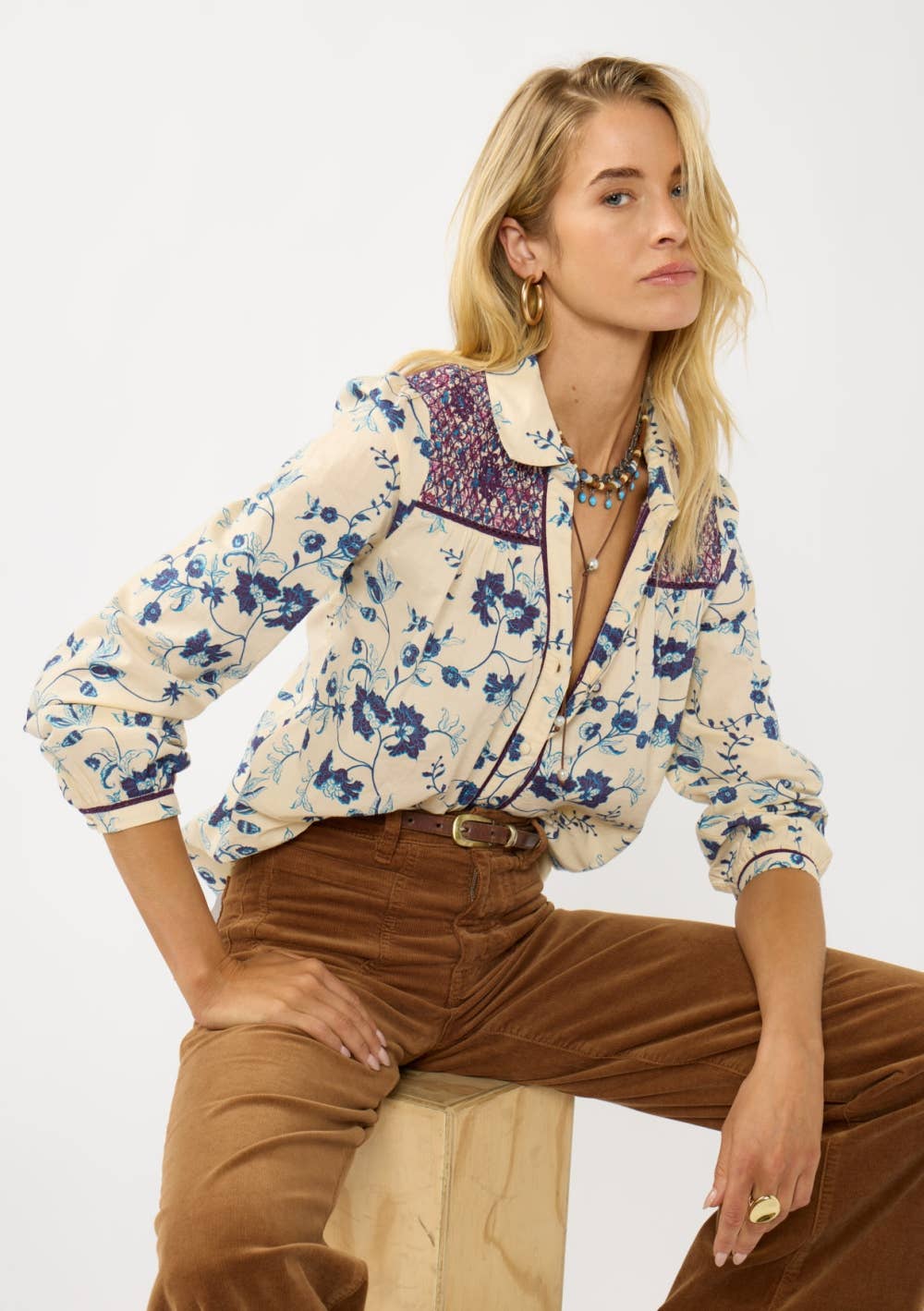 Woman wearing a Long Sleeve Floral Blouse with Smocked Yoke in cream with blue floral print and brown pants sitting on a stool