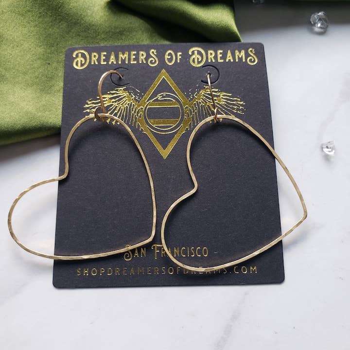 The Big Gold Heart Hoop Earrings by Dreamers of Dreams, shown on a branded black card with gold details, rest elegantly on marble beside a green cloth and crystals—perfect statement hoops for any look.