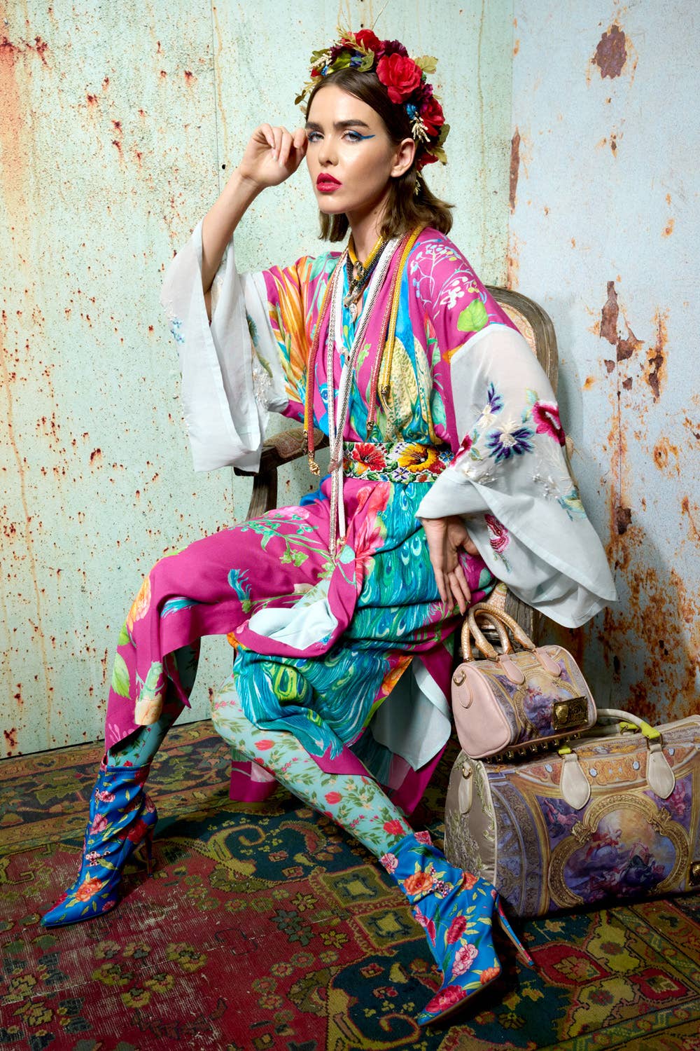 A woman in floral boots sits on a chair against a textured wall, wearing the Aratta Marie Antoinette Kimono as a statement piece. She completes her artistic look with a flower crown, bold makeup, and a decorated bag.