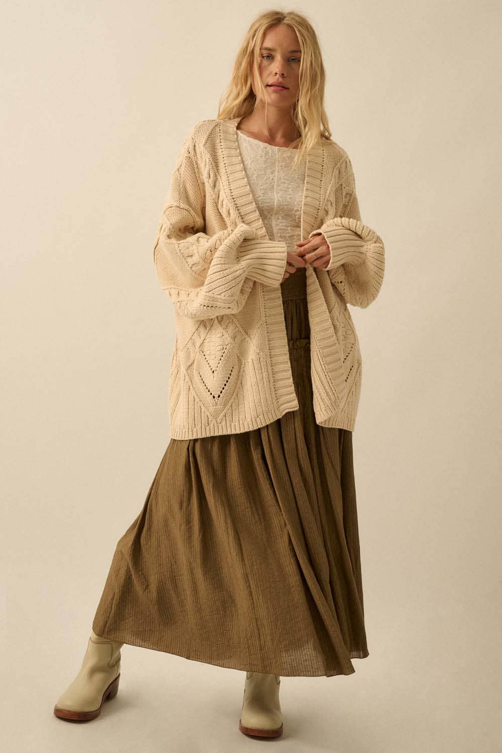 A woman with long, wavy blonde hair and a relaxed expression stands against a beige background, wearing the Promesa USA Cable-Knit Oversize Balloon-Sleeve Cardigan over a white top, an olive-green pleated skirt, and light ankle boots.