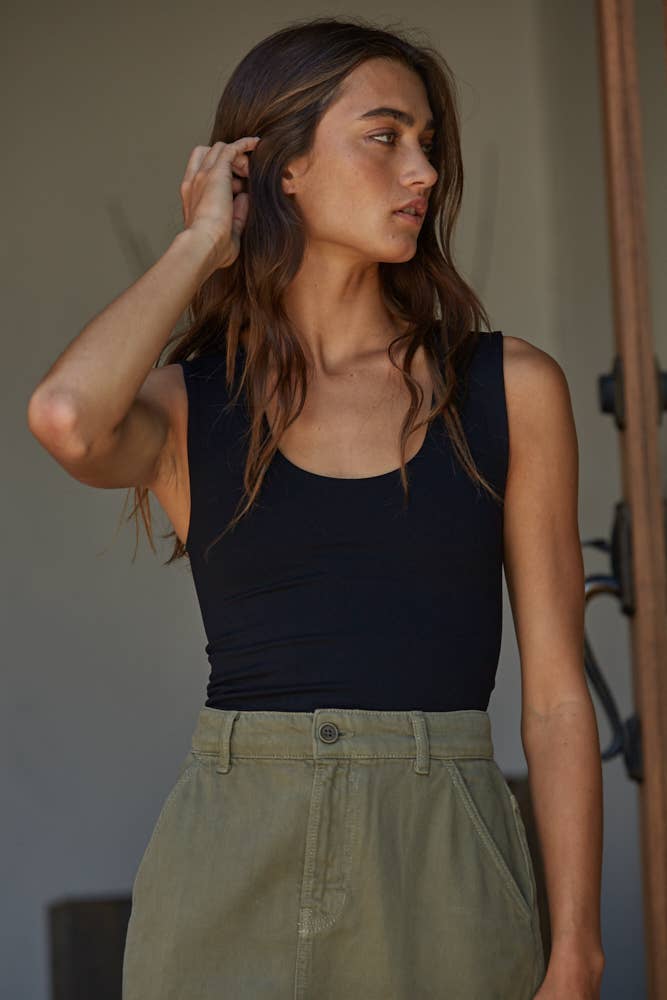 A woman with long brown hair stands indoors, touching her hair and looking left, wearing the By Together Double Layered Seamless Top and high-waisted olive green pants.