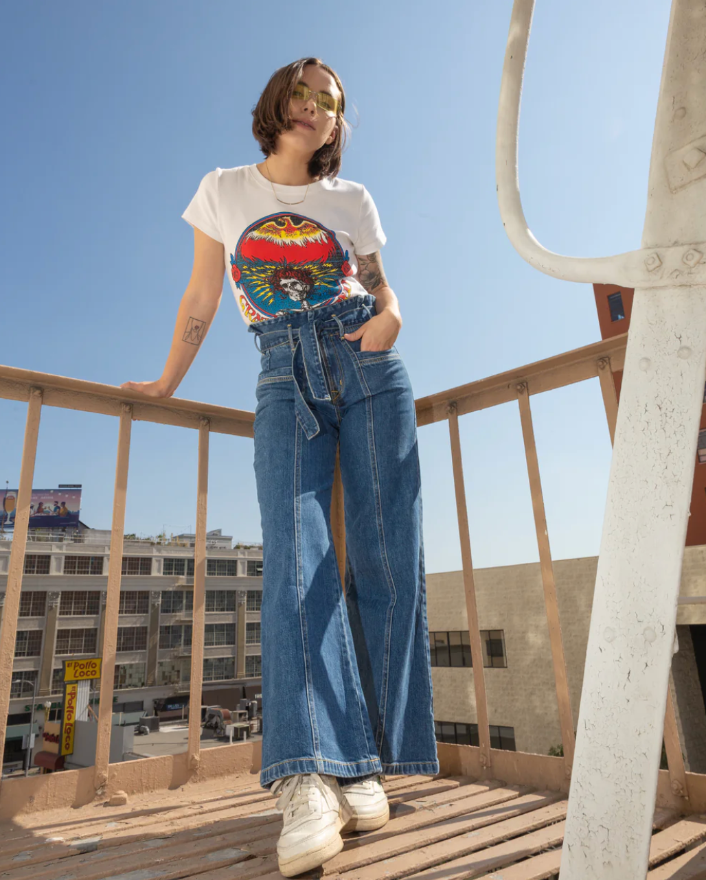 A woman in sunglasses, a graphic tee, and Porter Blue Apparel’s PENNY PAPERBAG WIDE LEG IN LAGUNA WASH jeans stands on an outdoor metal staircase, leaning on the railing with city buildings behind her under a clear sky.