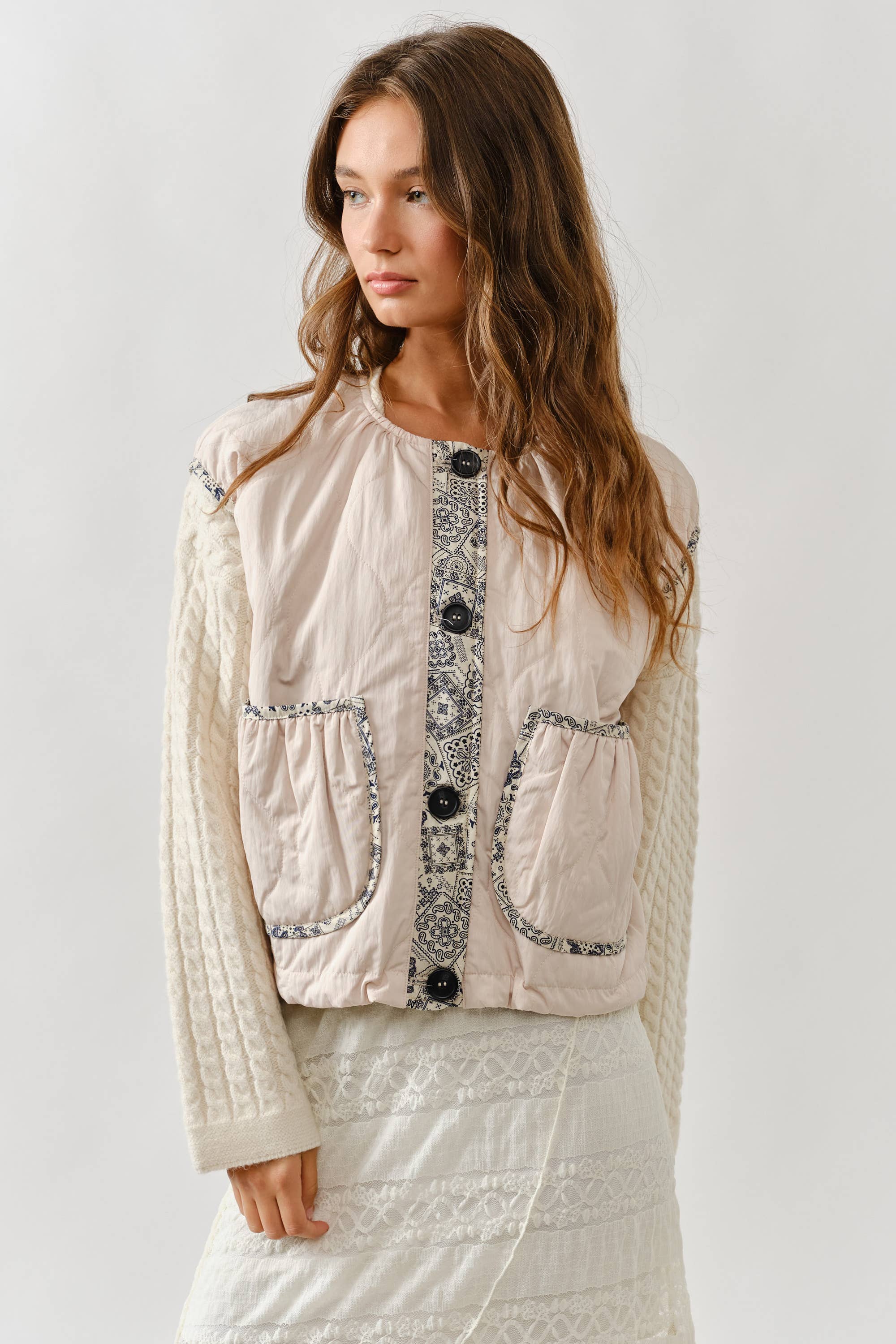 A woman with long brown hair wears a cream sweater, a white lace skirt, and the Aaron & Amber Contrast Detail Lightweight Vest—a layering essential with patterned trim—standing against a plain light background.