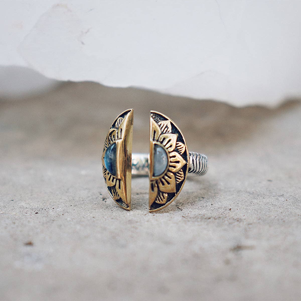 The Natural Mystic Ring - Moonstone by bahgsu jewels—a sterling silver adjustable ring with a blue moonstone, framed by gold-toned sunburst-engraved semicircles—is displayed on a neutral textured surface, perfect for any collection.