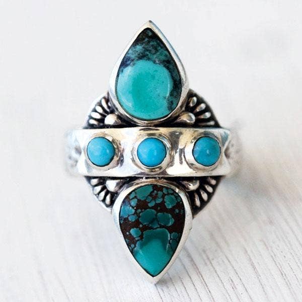 The bahgsu jewels Truth Seeker Ring features two teardrop-shaped turquoise stones, three small round gems, and ornate detailing. This silver ring is uniquely designed and beautifully displayed on a light surface.