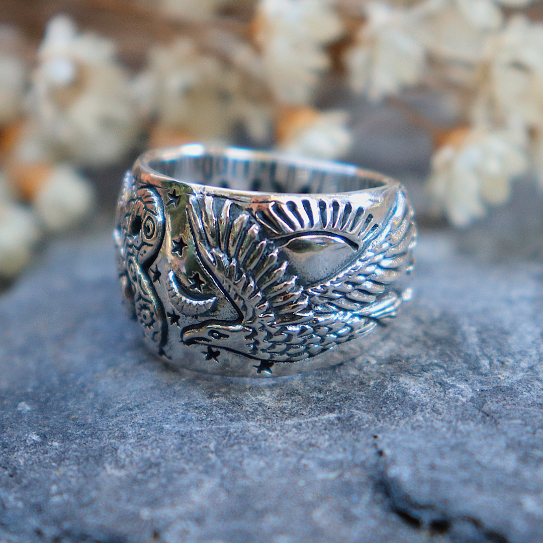 A symbolic piece, the Rainbow Serpent Ring by bahgsu jewels (Xavier Rudd Collab) features detailed engravings and rests on a gray stone surface with blurred white flowers in the background.