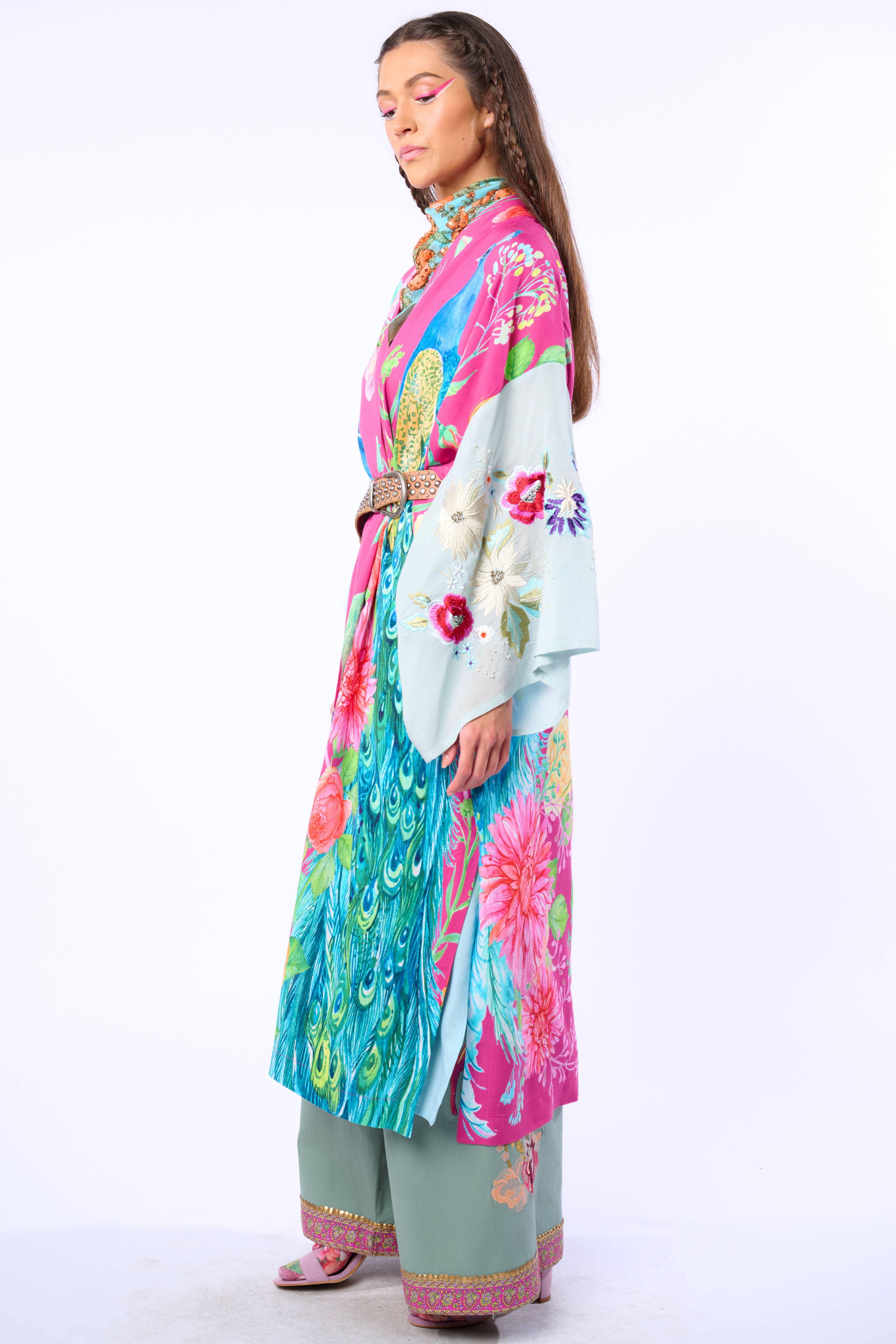 A woman stands sideways, eyes closed, wearing Aratta’s Marie Antoinette Kimono with embroidered floral sleeves and wide cuffs, layered over green pants with pink embroidery and matching sandals, set against a plain white background.