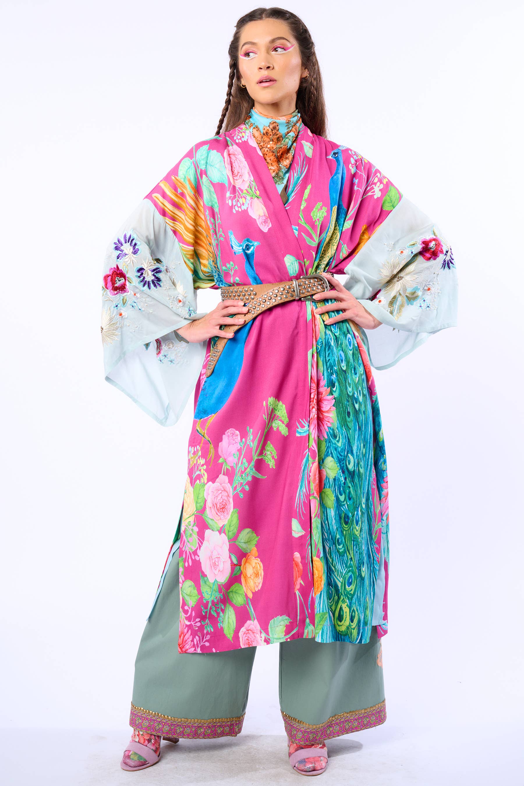 A woman exudes confidence in the Aratta Marie Antoinette Kimono with embroidered floral sleeves, styled with sage green pants, a decorative belt, pink shoes, and bold makeup, all set against a plain white background.