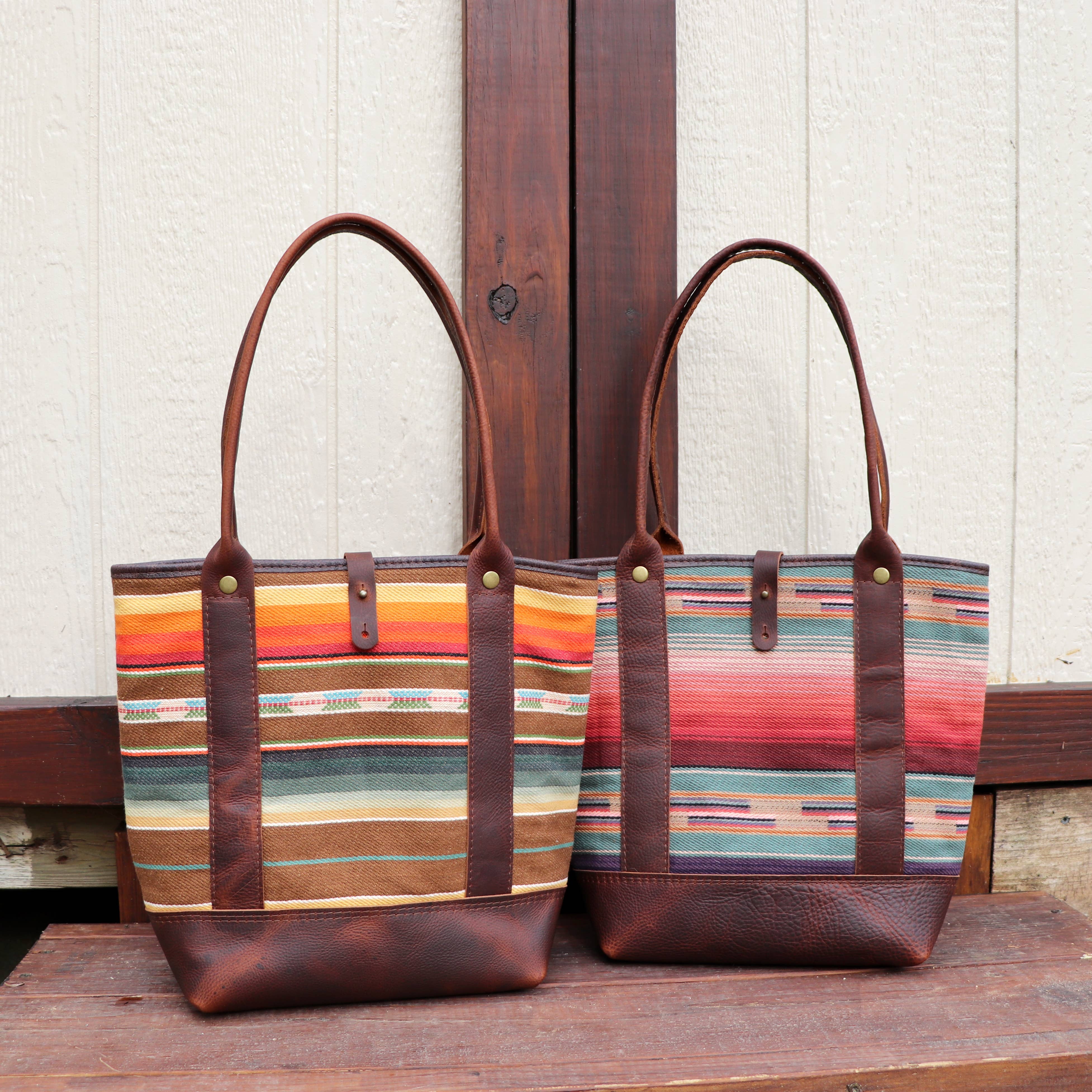 Two Pearl Serape Totes from Hawks & Doves, each with colorful serape fabric in earthy and vibrant stripes, full grain leather handles and bottoms, are displayed side by side against a wooden background.