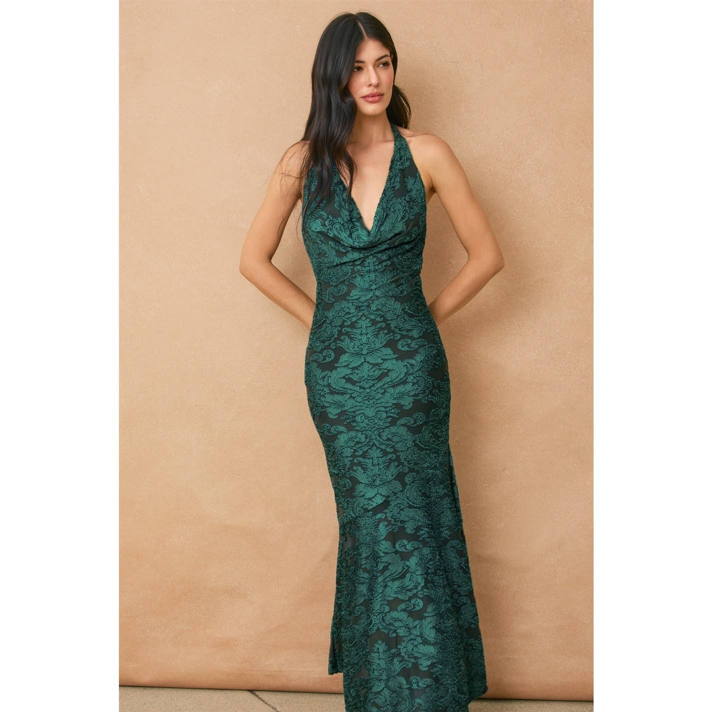 A woman with long dark hair wears the Fame Draped Halter Neck Maxi Dress by Dress Forum, a sleeveless emerald green floral mesh design, standing against a beige background.