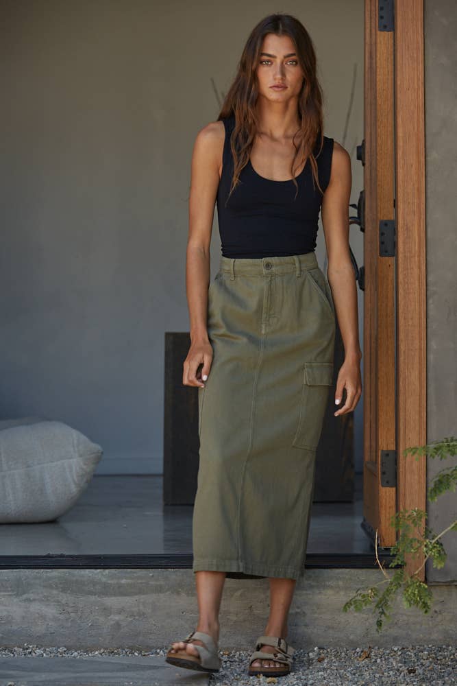 A woman with long brown hair and a neutral expression stands in a modern, minimalistic doorway wearing the By Together Double Layered Seamless Top, an olive green cargo midi skirt, and sandals.
