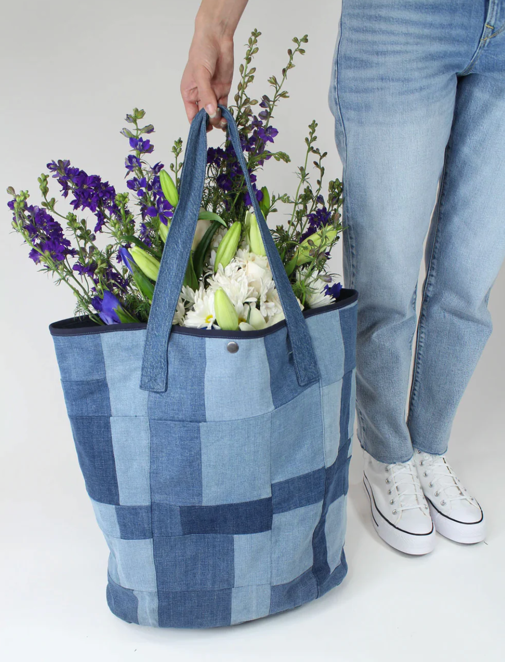 A person in blue jeans and white sneakers holds the Porter Blue Apparel PATCHWORK TOTE filled with purple and white flowers.