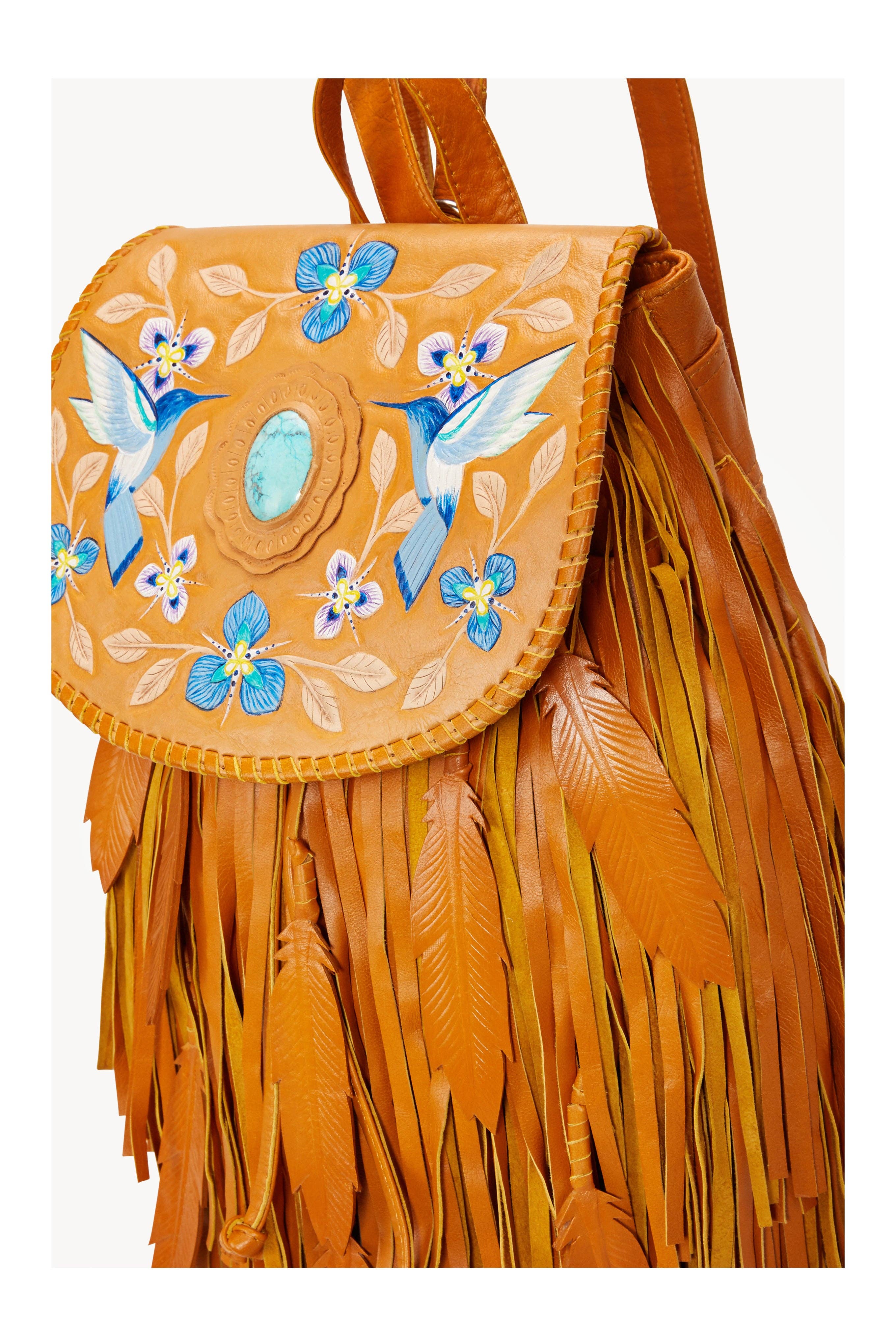 The Monterey Backpack in Antique Tan by Jodi Lee is a handmade leather bag with long fringe, embossed feathers, hand-painted blue flowers and birds, and a turquoise-style stone accent on the flap.