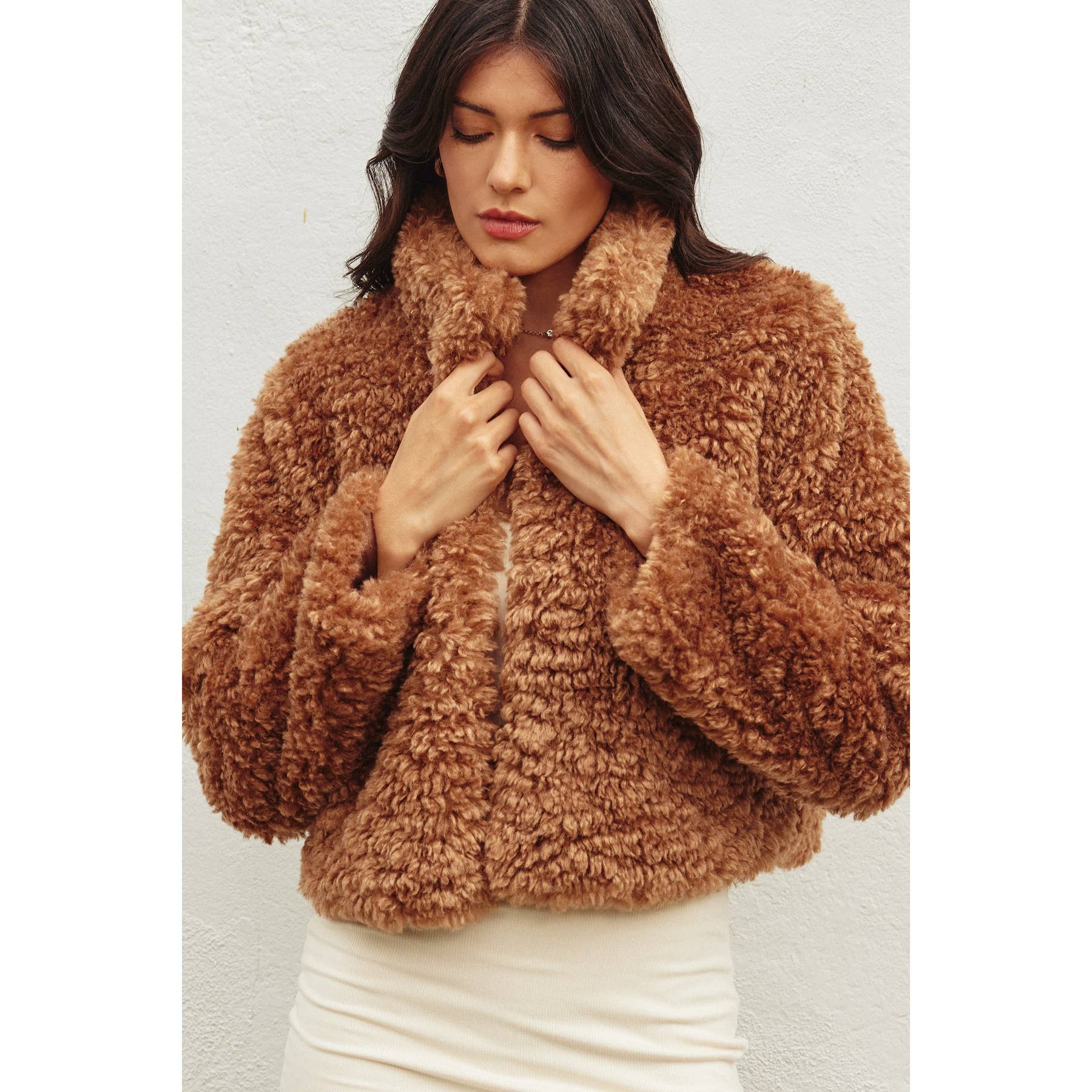 Woman with long dark hair wearing the Dress Forum Soft Teddy Open Front Cropped Jacket in brown over a light outfit, standing against a plain white background and looking down.