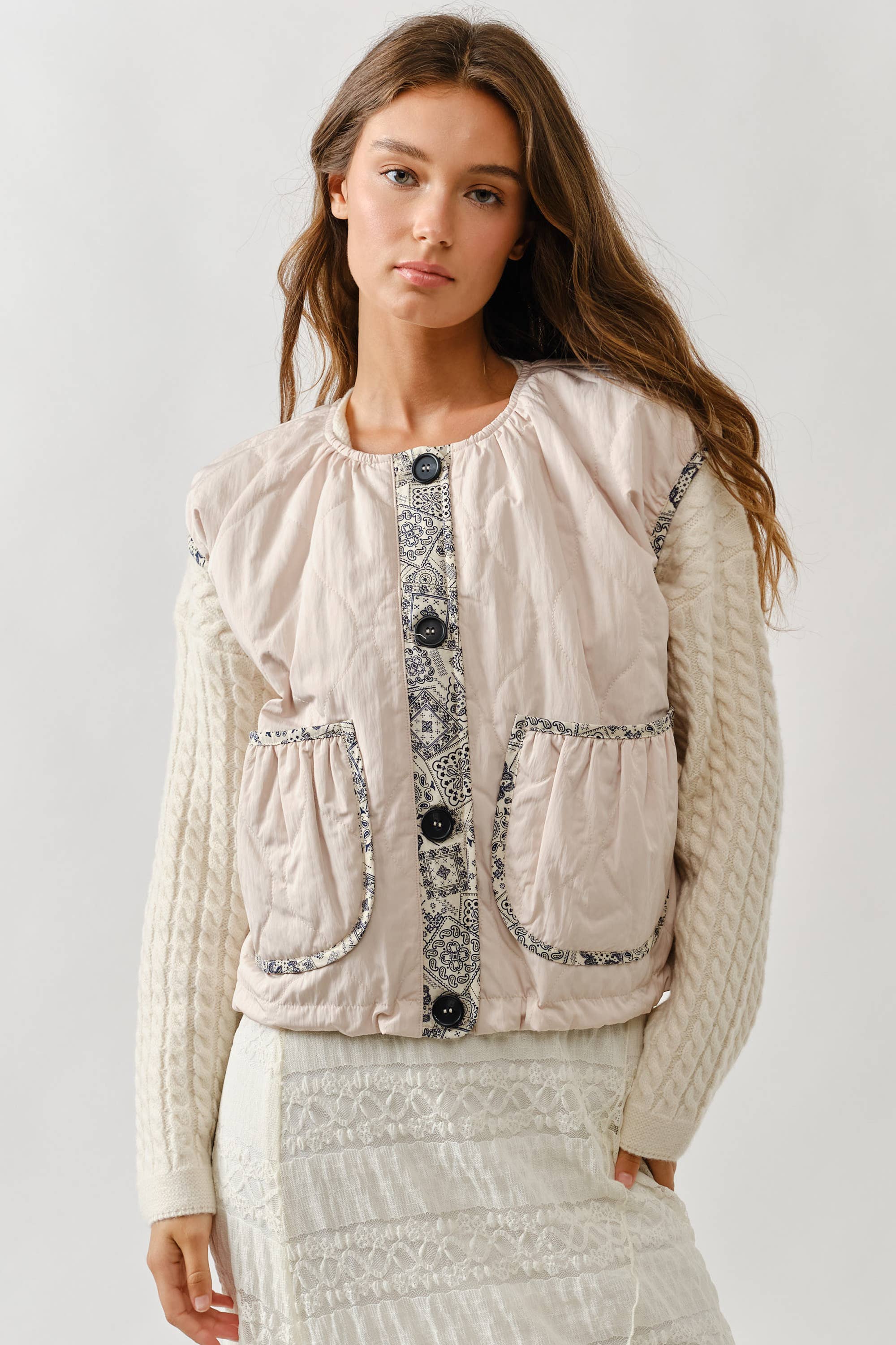 A woman with long brown hair wears the Aaron & Amber Contrast Detail Lightweight Vest—a pink quilted layering essential with patterned trim and large pockets—over a cream cable-knit sweater and skirt, standing against a plain light background.