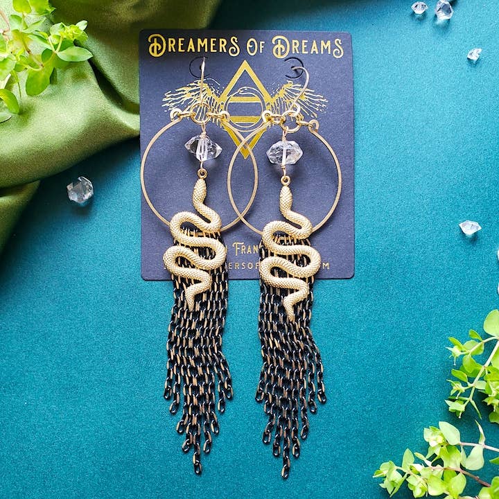 The Rising Snake Earrings by Dreamers of Dreams feature coiled gold designs and long black chain fringe, showcased on a branded card among green leaves and crystals—a bold, hypoallergenic accessory.