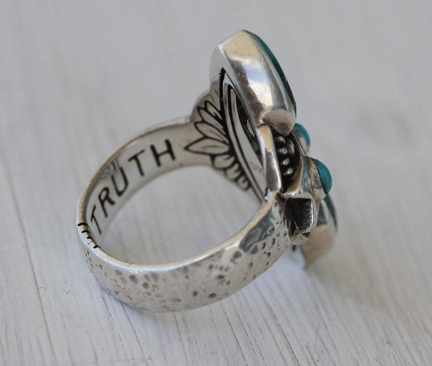 A close-up of the bahgsu jewels Truth Seeker Ring—featuring engraved flowers, turquoise stones, and TRUTH etched inside—photographed on light wood for a mindful, unique style.