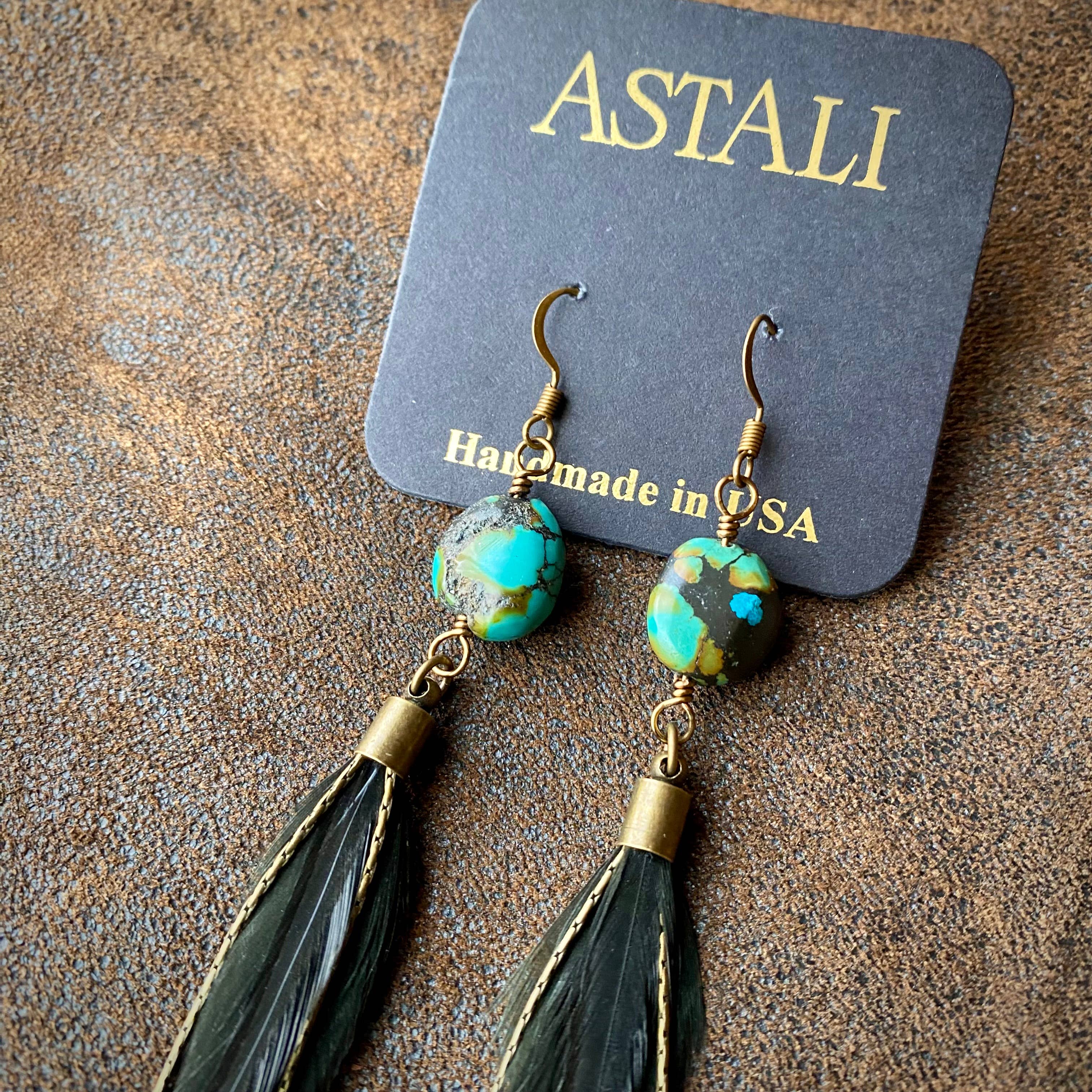 ASTALI Feather & Turquoise Statement Earrings feature antique brass and sustainable coque feathers, accented with turquoise stones and displayed on a branded card against a textured brown background. Handmade in the USA.