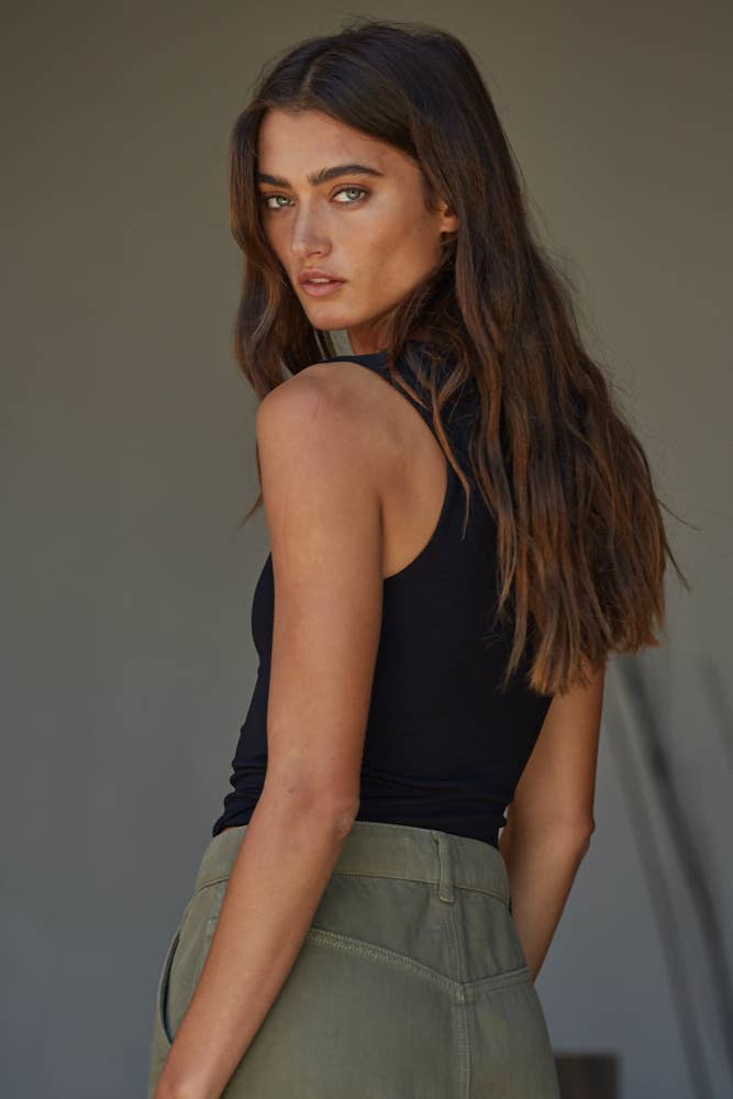 A woman with long brown hair wears a By Together Double Layered Seamless Top and green pants, looking over her shoulder at the camera against a plain, neutral background.