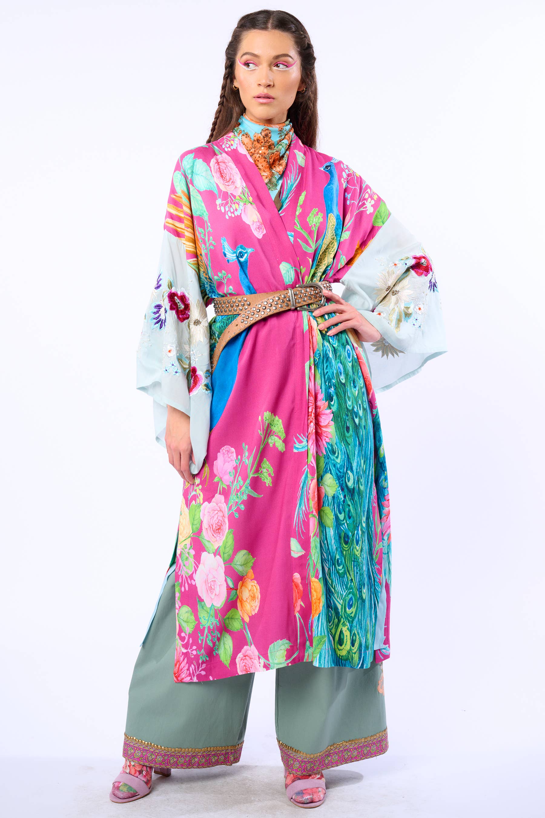 A woman stands confidently against a white background in the Aratta Marie Antoinette Kimono—featuring embroidered floral sleeves—styled with a woven belt, loose green pants, and pink sandals.