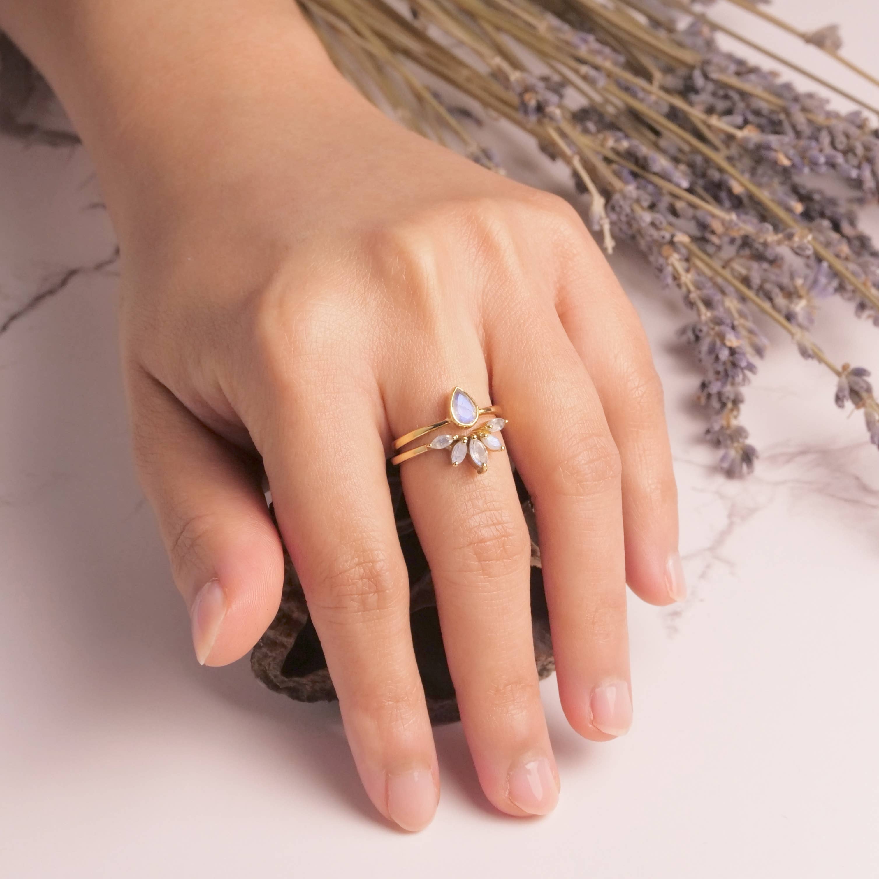 A hand with neatly manicured nails wears the Baizaar Brass Nesting Moonstone Ring Set, resting on a white marble surface beside dried lavender sprigs.