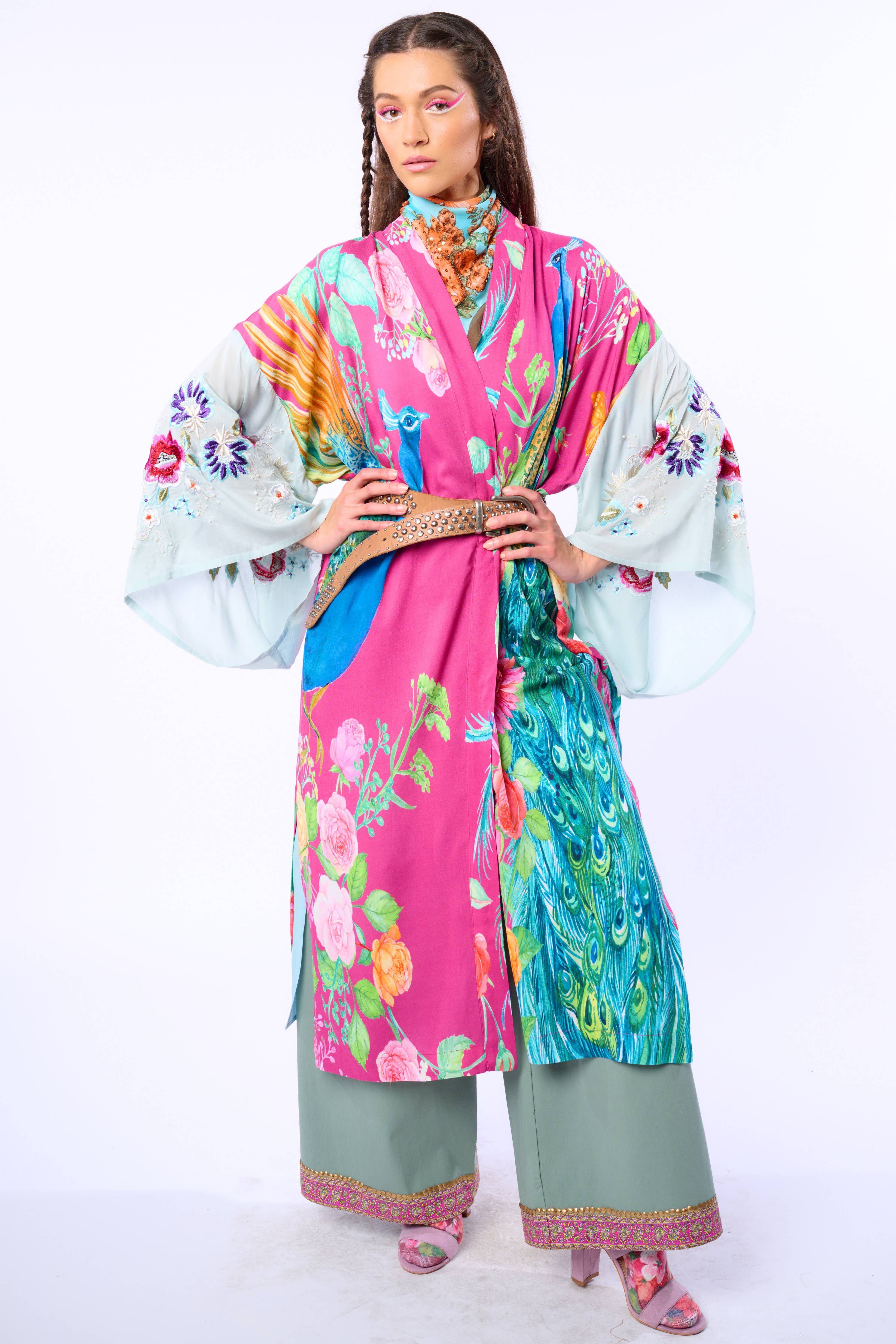 A person poses confidently in the Aratta Marie Antoinette Kimono, featuring embroidered floral sleeves and wide cuts, paired with loose green pants and pink heels—creating a striking statement look against a white background.