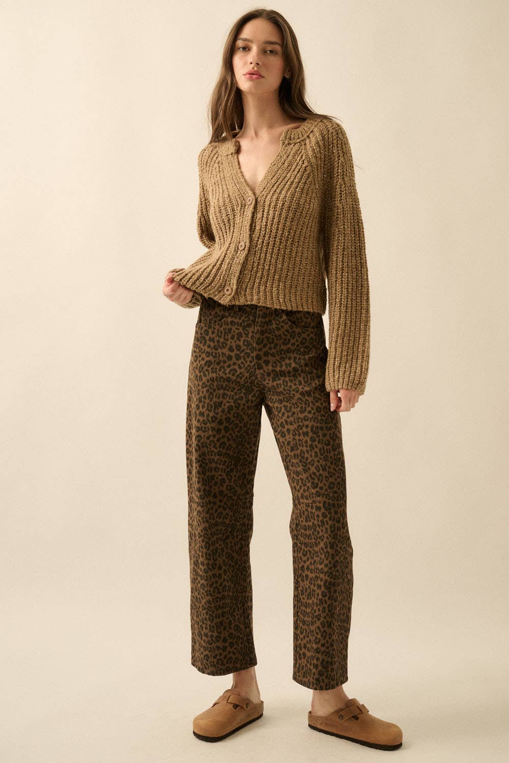 A woman with long brown hair and a neutral expression stands against a beige background wearing the Promesa USA Confetti Button-Front Cardigan Sweater, paired with brown leopard-print pants and tan slip-on shoes, one hand holding her sweaters edge.