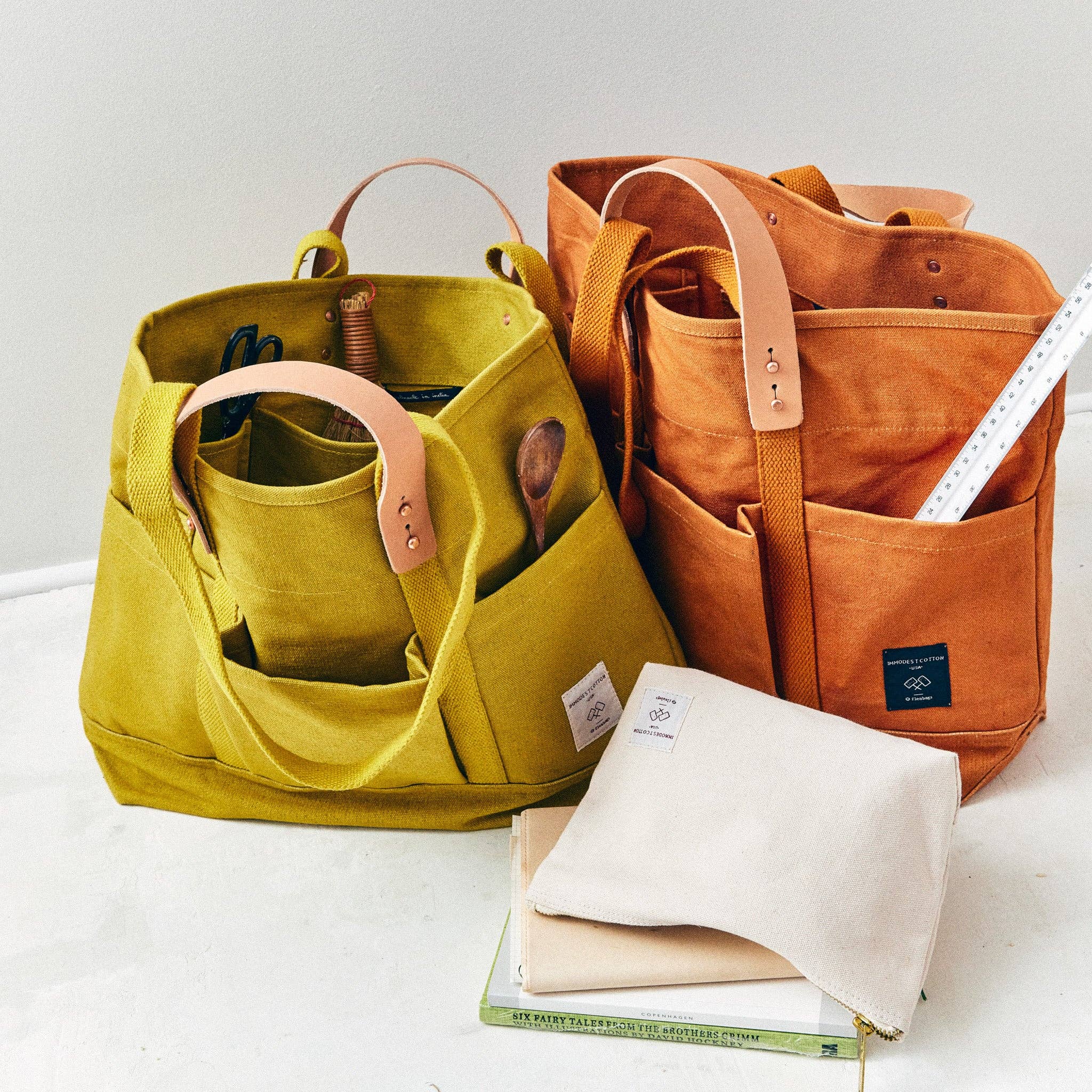 Two sturdy canvas bags with leather handles in mustard seed and rust hues filled with tools and accessories, including Construction Tote | Mustard Seed